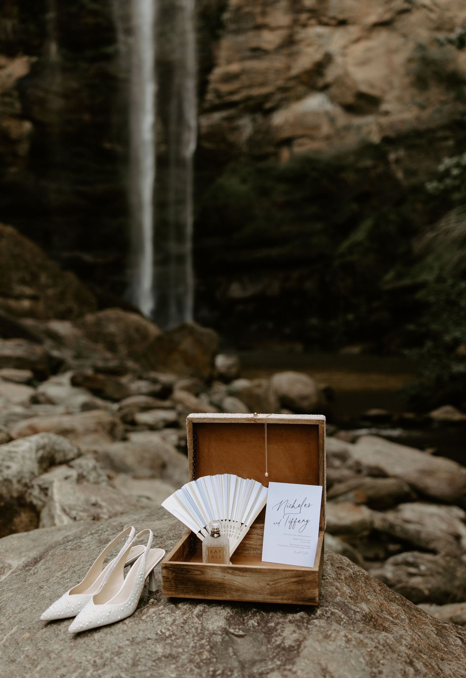 Elegant white wedding accessories including pointed shoes and cards arranged on rustic stone surface against waterfall backdrop.