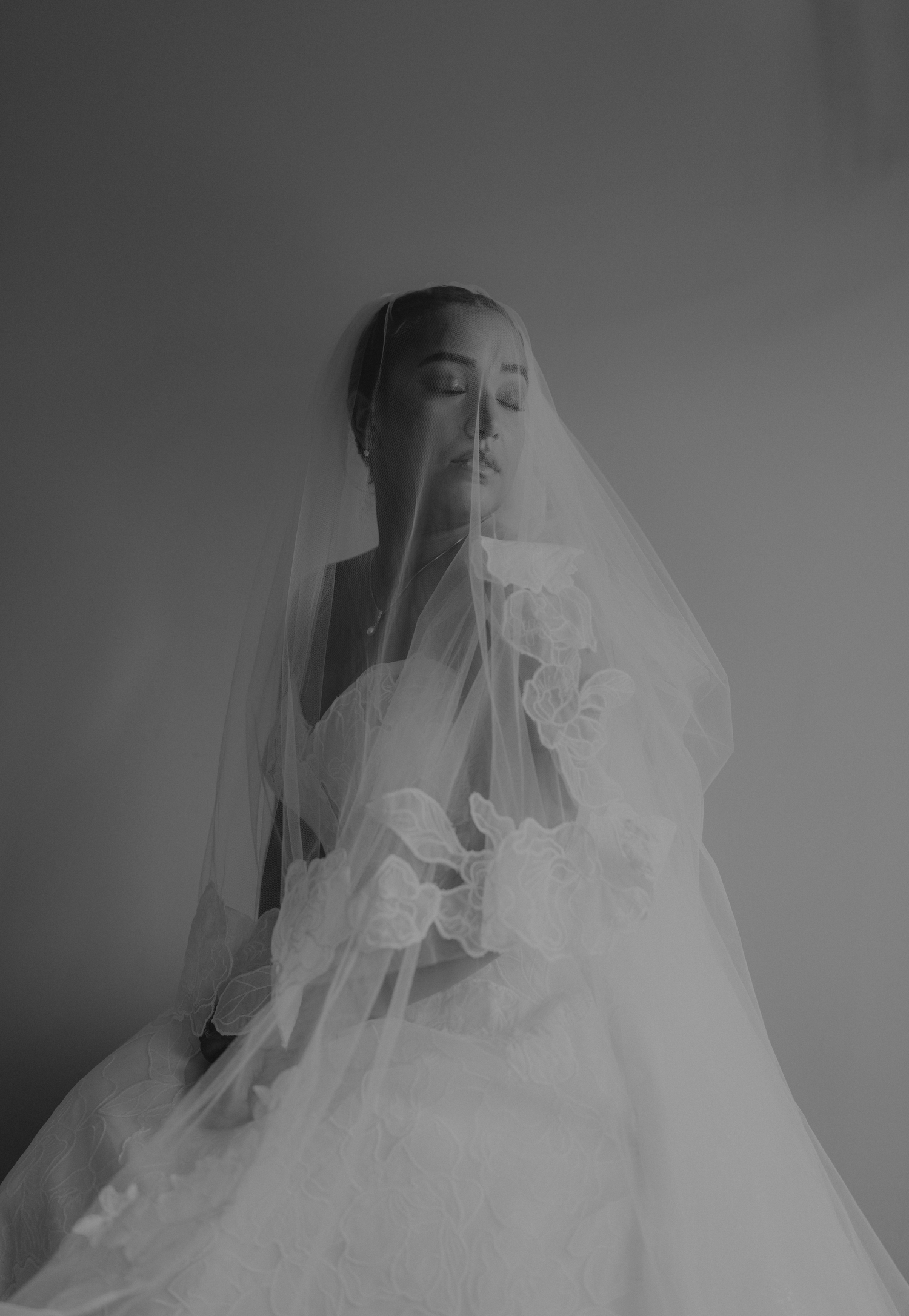 Bride in a white wedding gown and floral veil, eyes closed, posing gracefully in a black and white portrait.