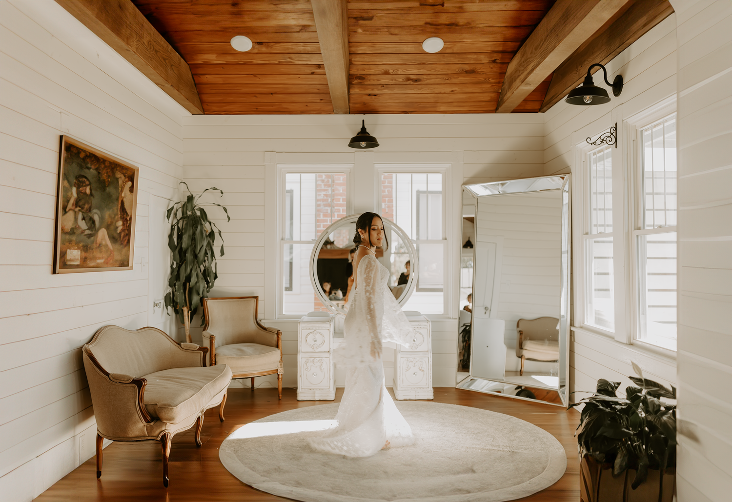 Elegant sunlit room with wood beam ceiling, white walls, round rug, vintage furniture and potted plants in a boho style interior.