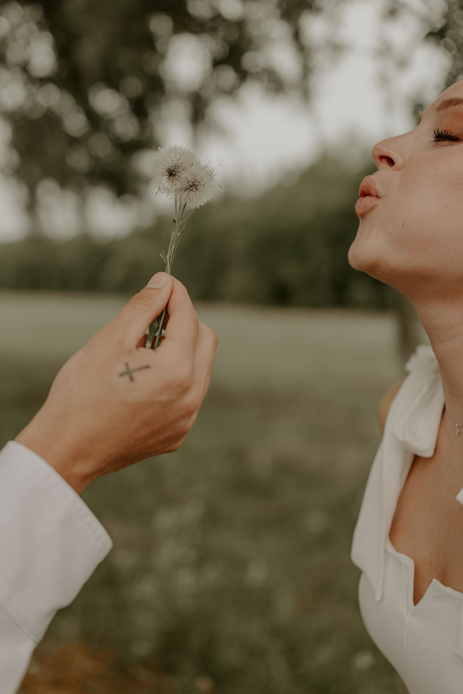 A person in a white top holds up and blows on a delicate dandelion seed head in dramatic natural lighting.