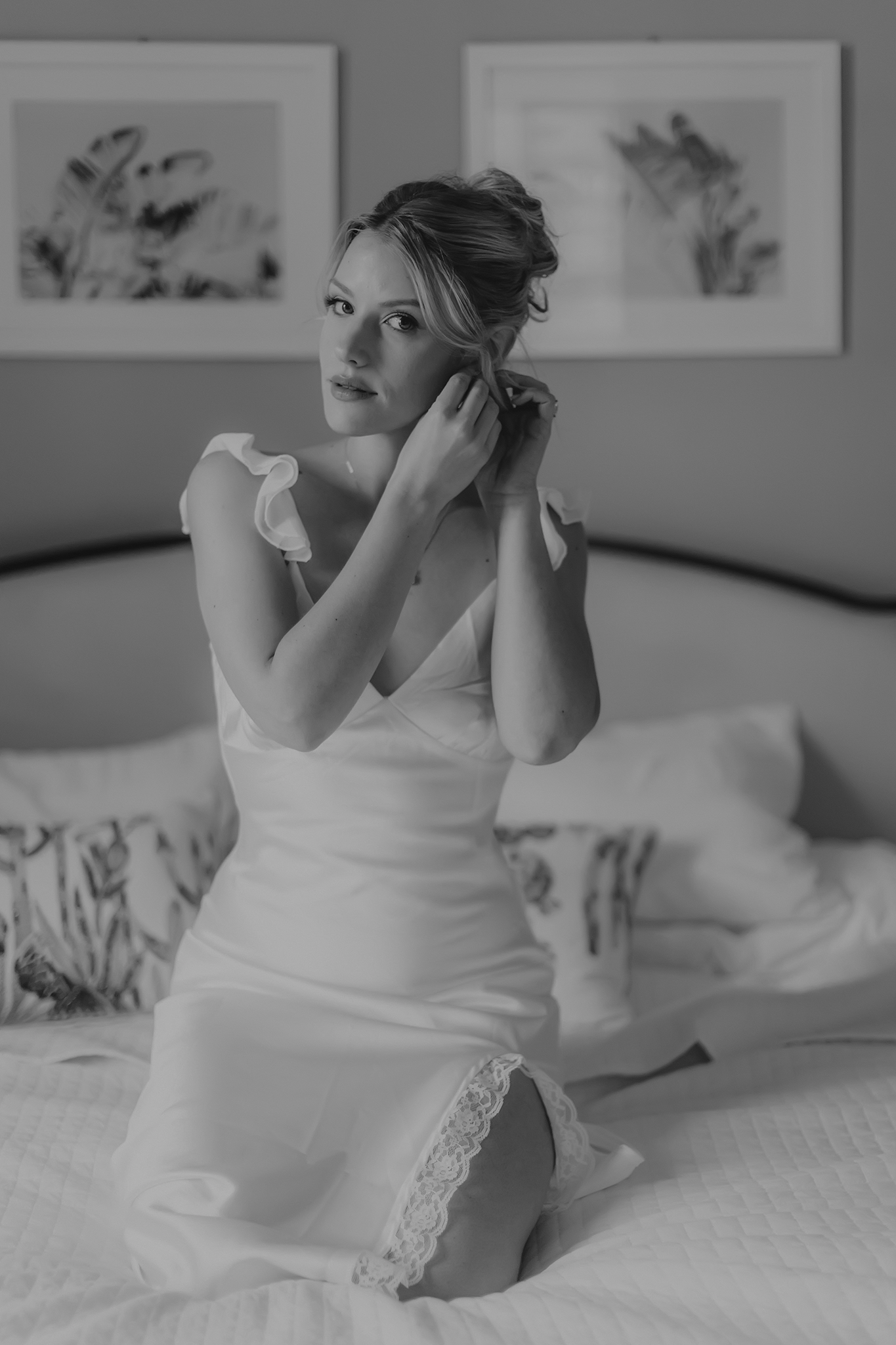 A person in a white dress reclines gracefully across a bed, creating an intimate boudoir scene in black and white.