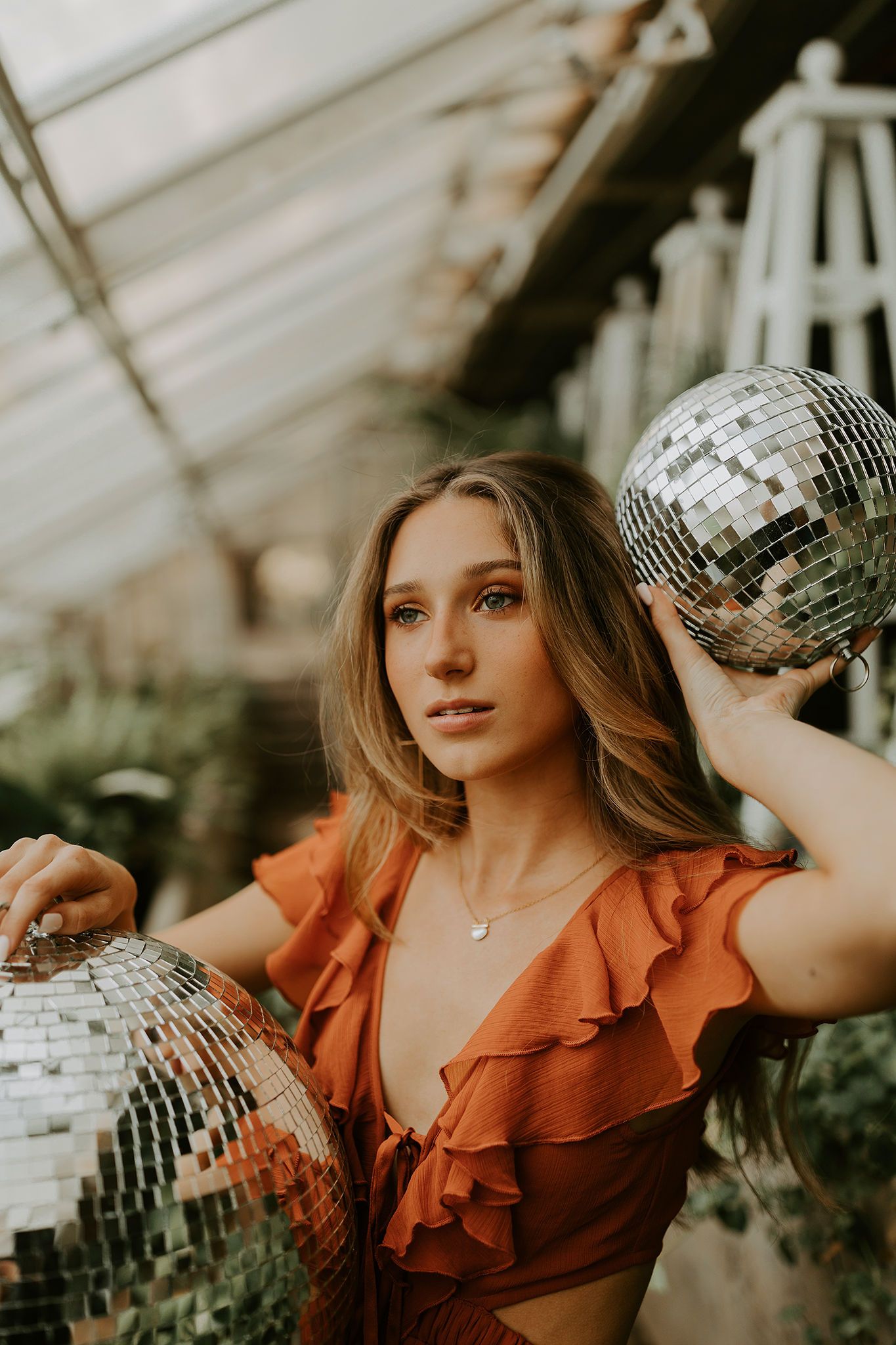 Person in rust-colored ruffled dress poses with disco balls in greenhouse setting during golden hour.