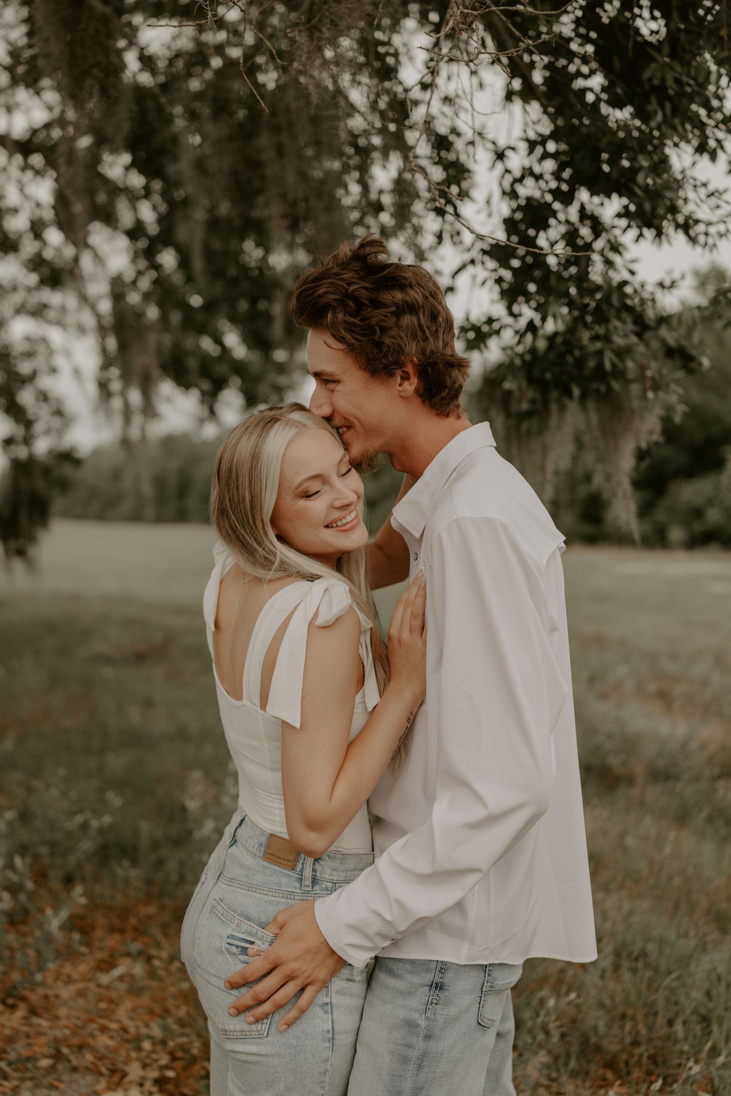 Romantic couple embraces outdoors in warm sunlight under mossy oak trees creating intimate engagement moment.