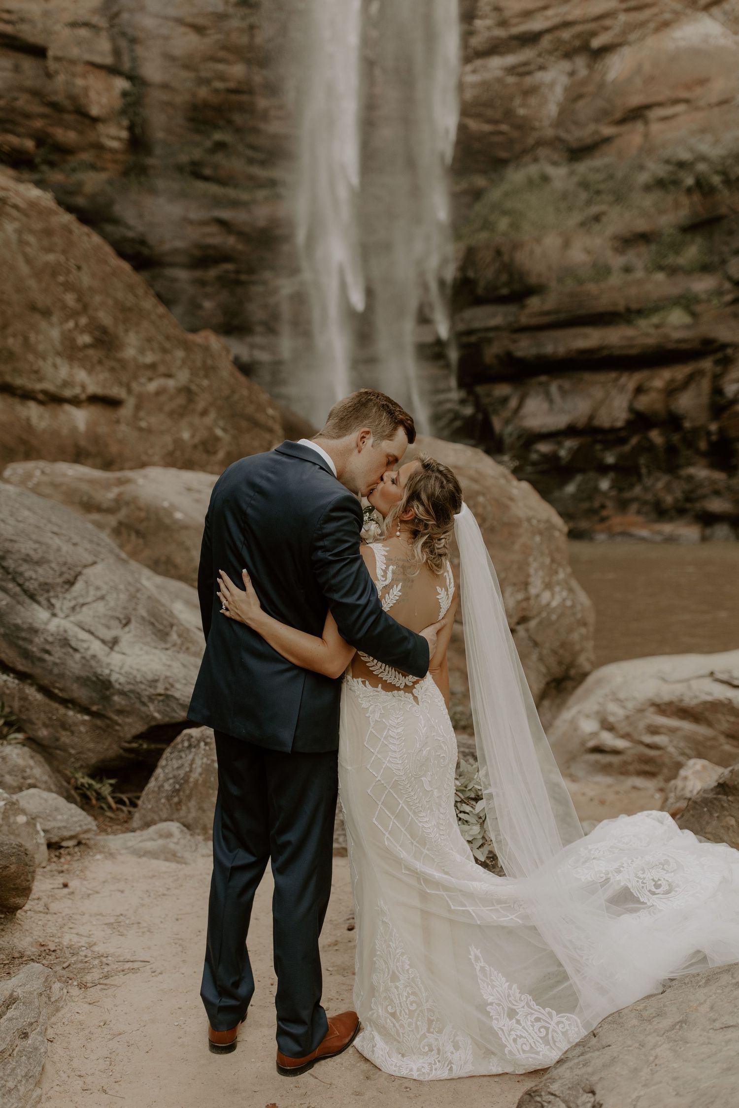 Newlyweds share an intimate moment embracing beneath a majestic waterfall in a scenic canyon setting.