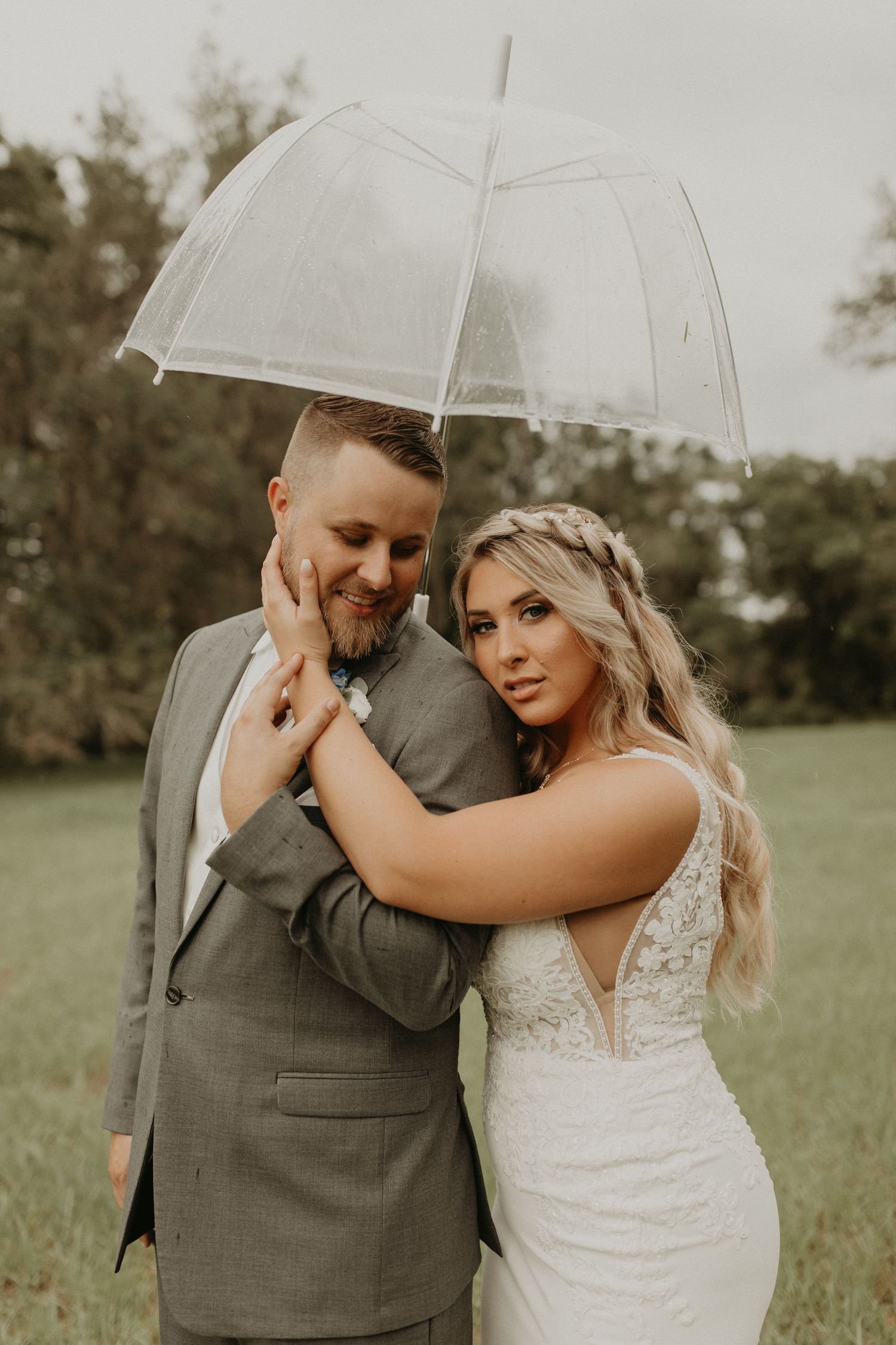 Romantic wedding portrait of a couple under a clear umbrella in a garden setting, wearing gray suit and white lace gown.