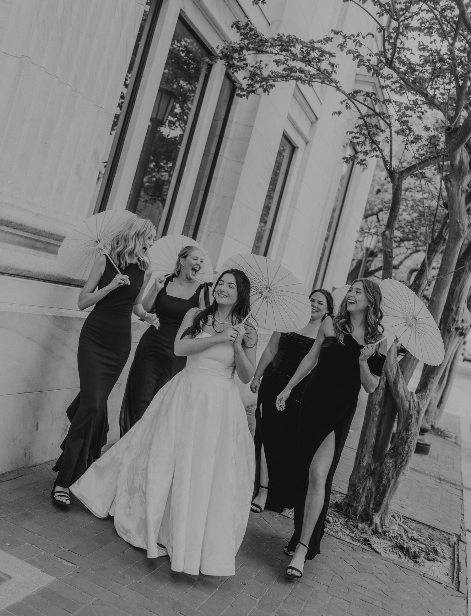 Bride in white gown laughs with bridesmaids in black dresses holding parasols outdoors in black and white photo.