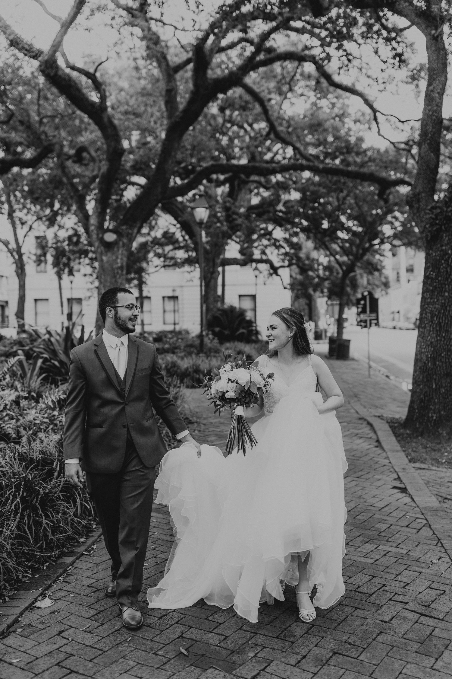 Black and white photo of a couple walking on a brick path under Spanish moss covered trees in wedding attire.