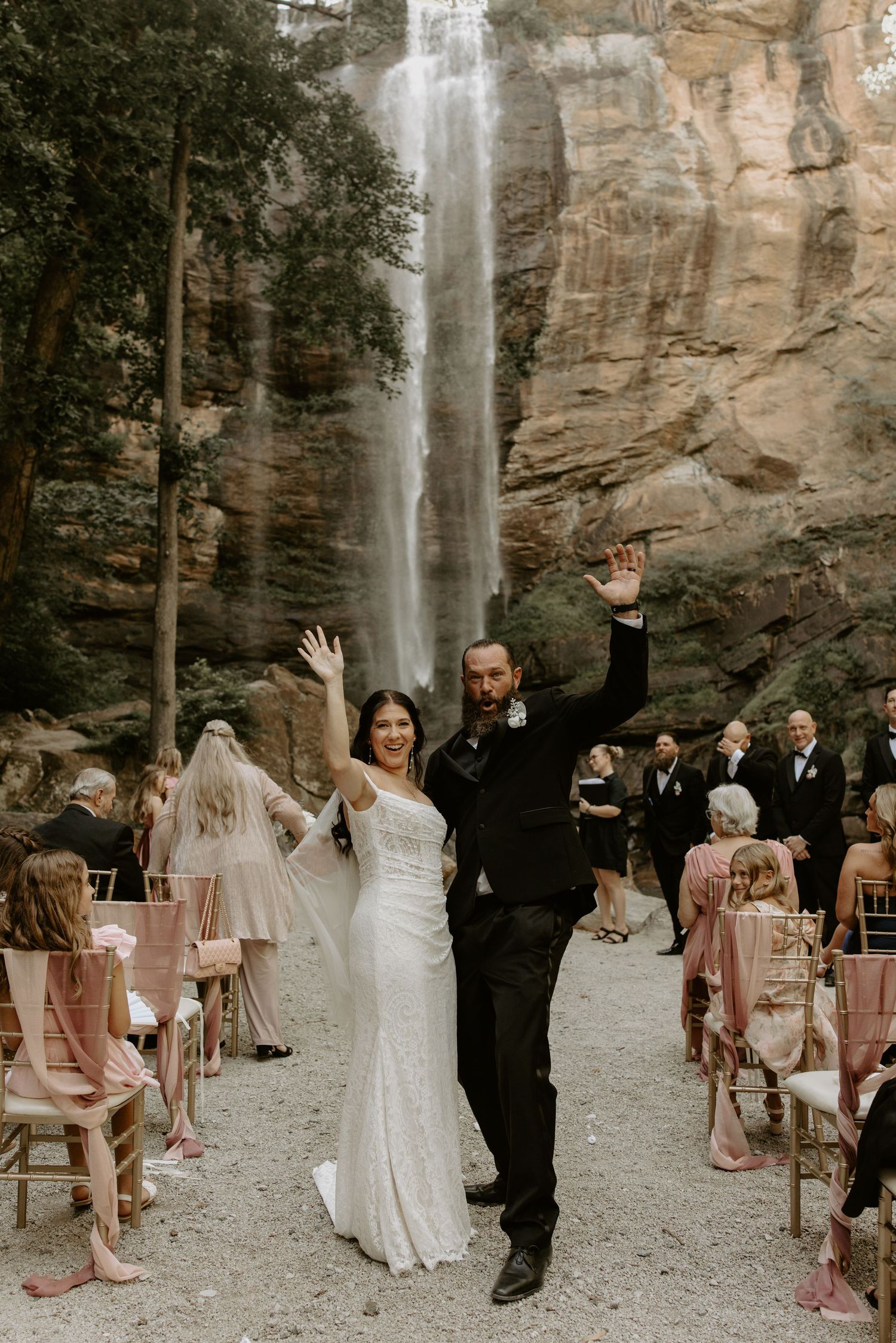 Intimate outdoor wedding ceremony takes place against dramatic backdrop of towering waterfall cascading down rocky cliffs.