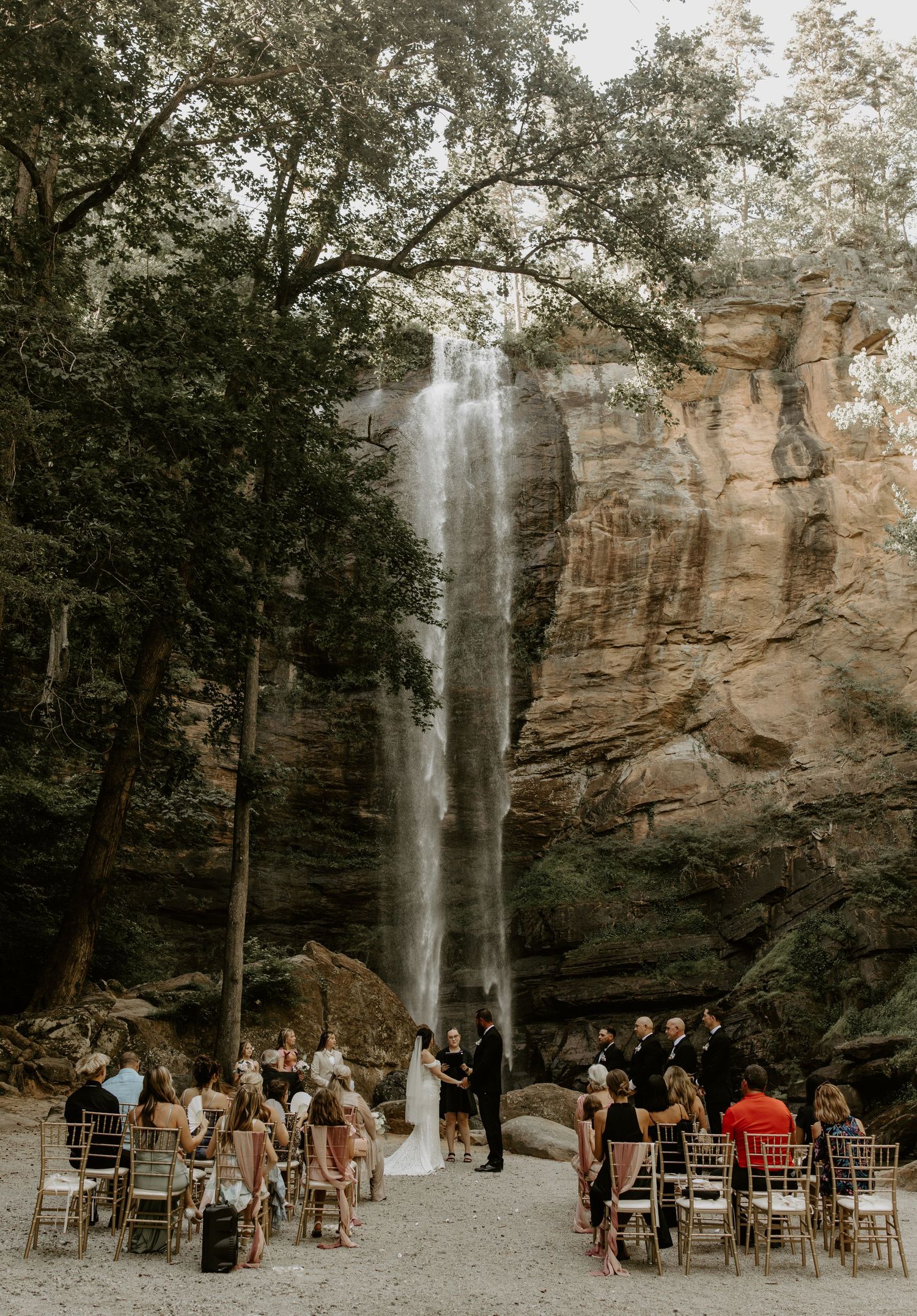 An outdoor wedding ceremony setup with wooden chairs arranged in front of a towering natural waterfall and rocky cliffs.