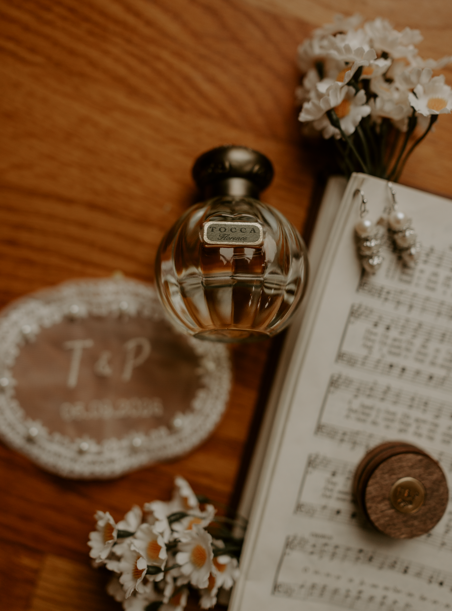 A vintage perfume bottle beside sheet music, daisies, and a monogrammed T&P wedding coaster on a wooden surface.