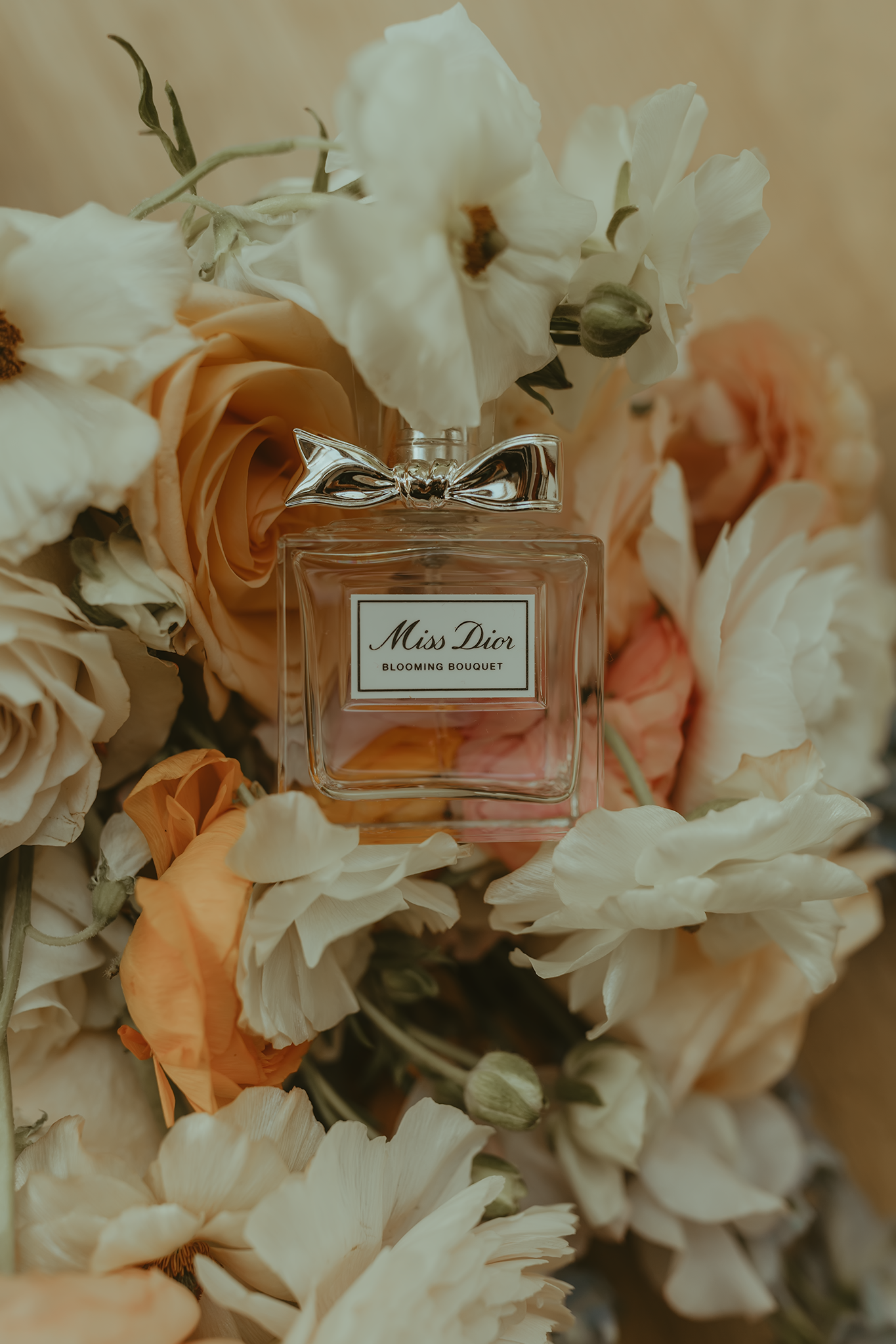 A bottle of Miss Dior perfume nestled among soft peach roses and white flowers in a dreamy vintage composition.