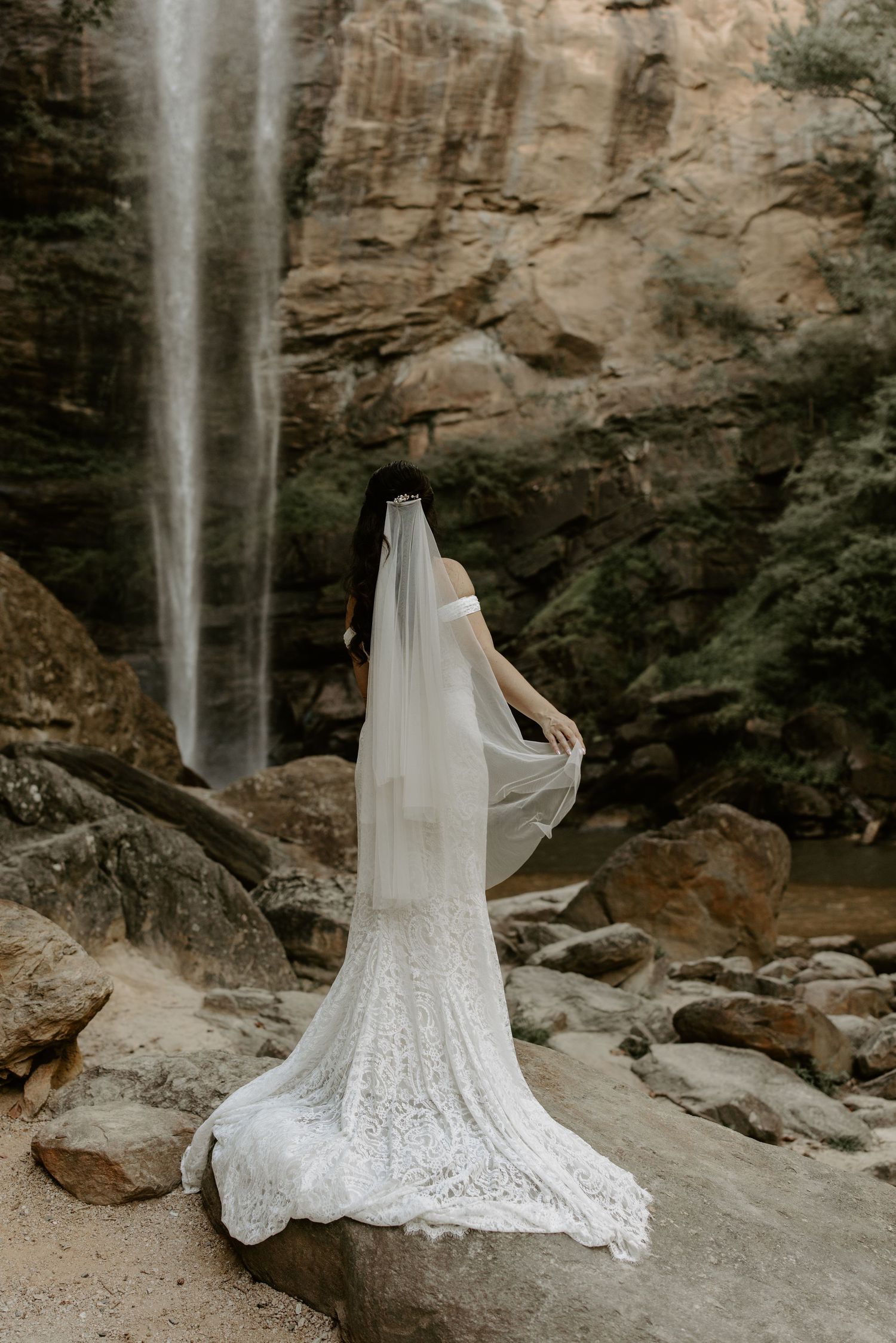 A flowing white wedding gown with intricate lace detailing photographed in front of a scenic waterfall against rocky cliffs.