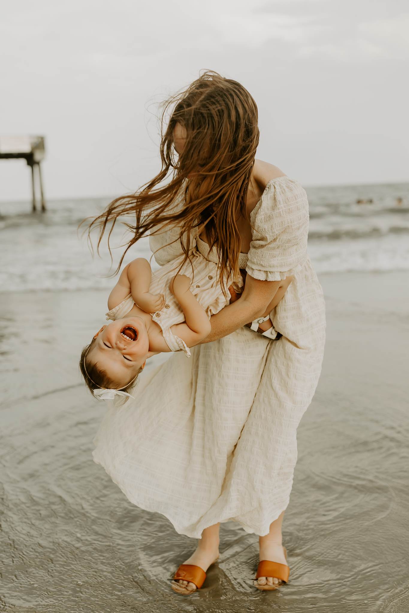 A candid moment between parent and child playing together on a beach in white dress and flowing hair.