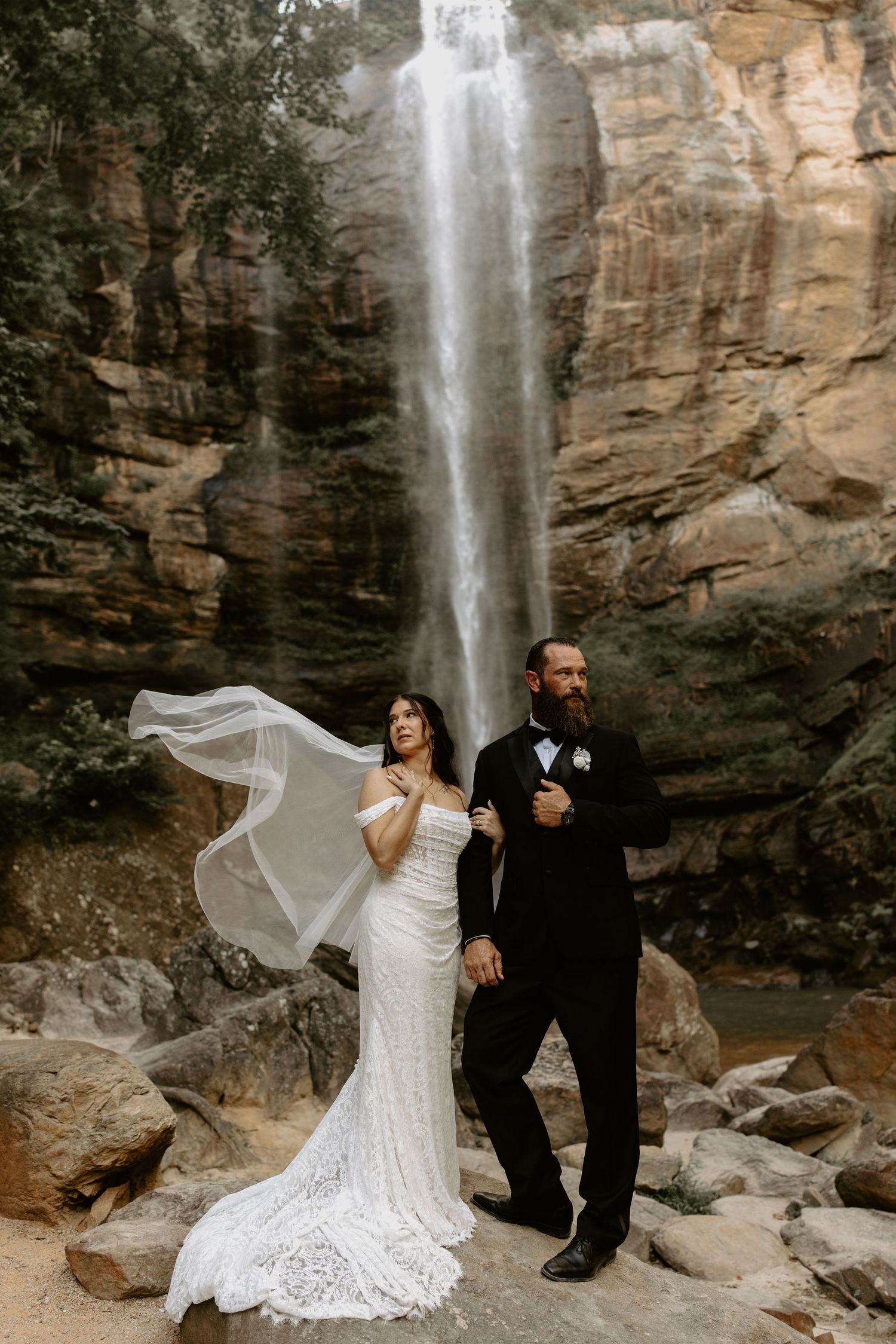 A couple poses at the base of a majestic waterfall with dramatic rock cliffs and flowing white veil trailing in the breeze.