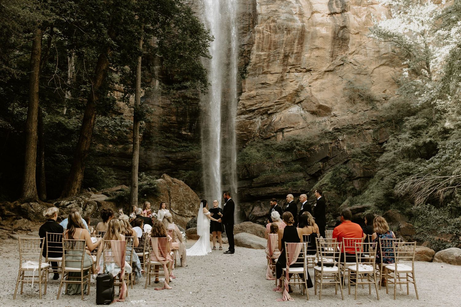 Elegant ceremony setup with wooden chairs arranged in rows facing a majestic waterfall cascading down tall rocky cliffs.