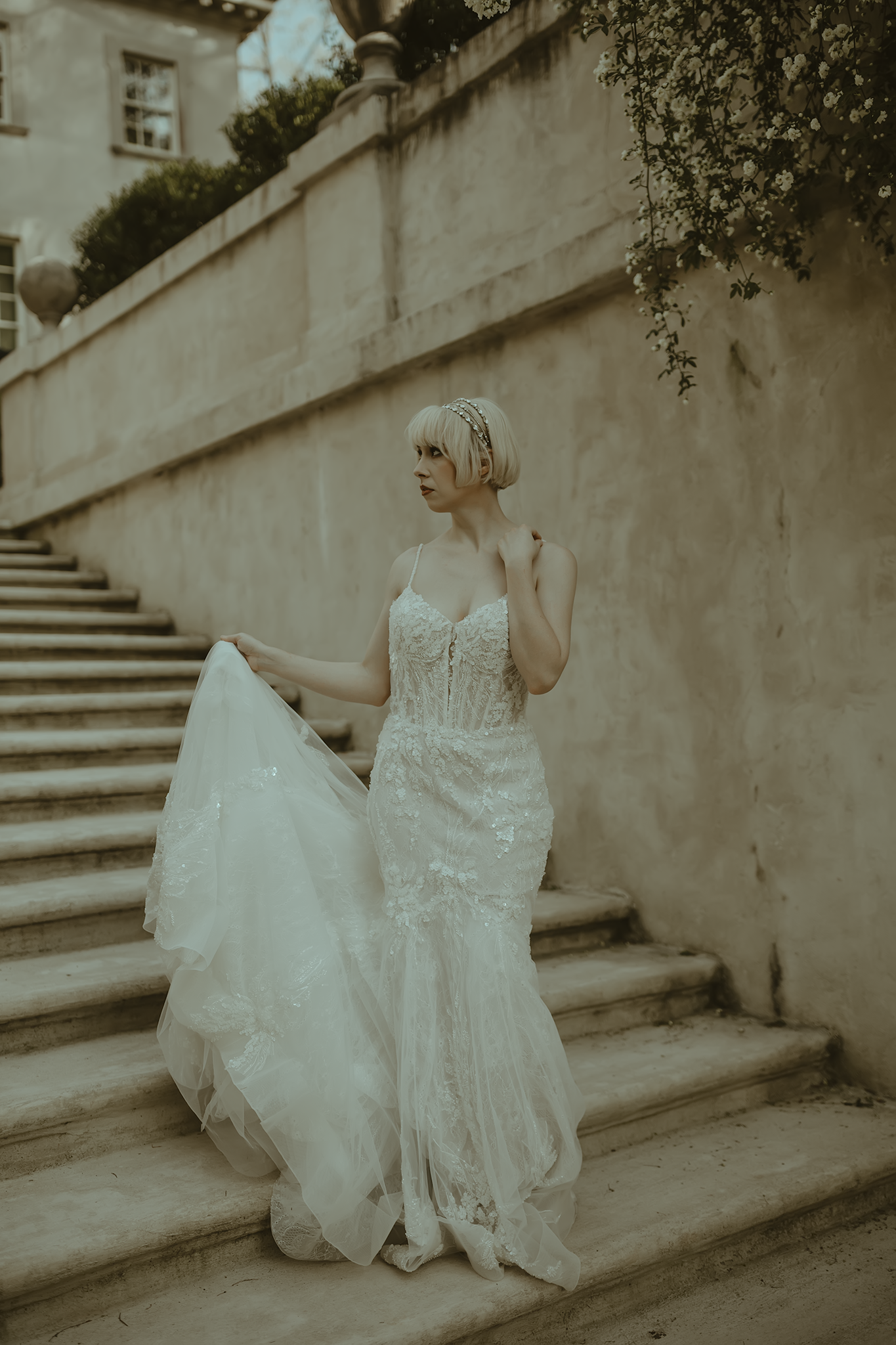 Bride in a vintage-style lace mermaid wedding gown poses elegantly on stone steps in a sepia-toned setting.