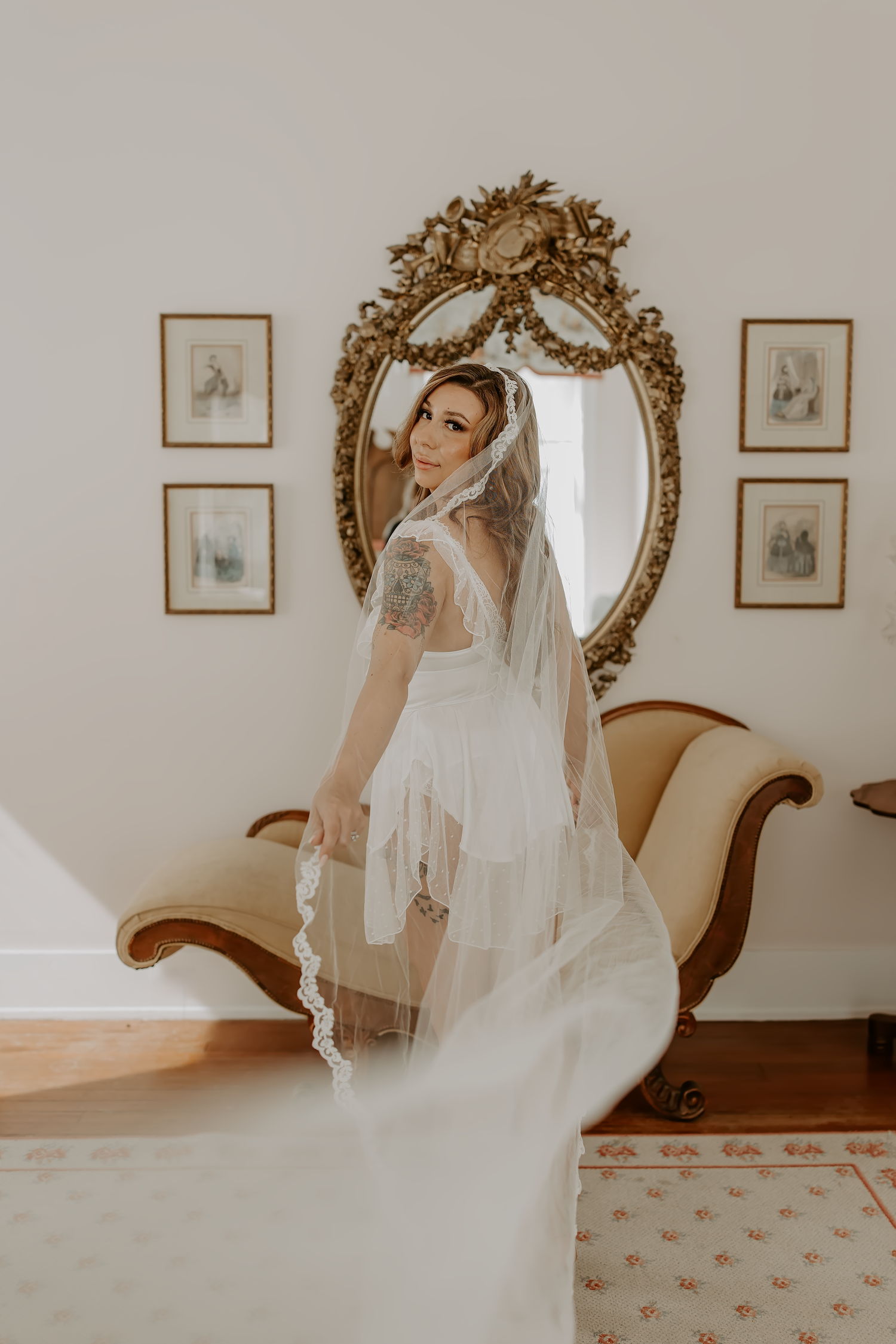 A bride in a flowing white wedding dress and veil poses elegantly in front of an ornate gold mirror with vintage frames on the wall.