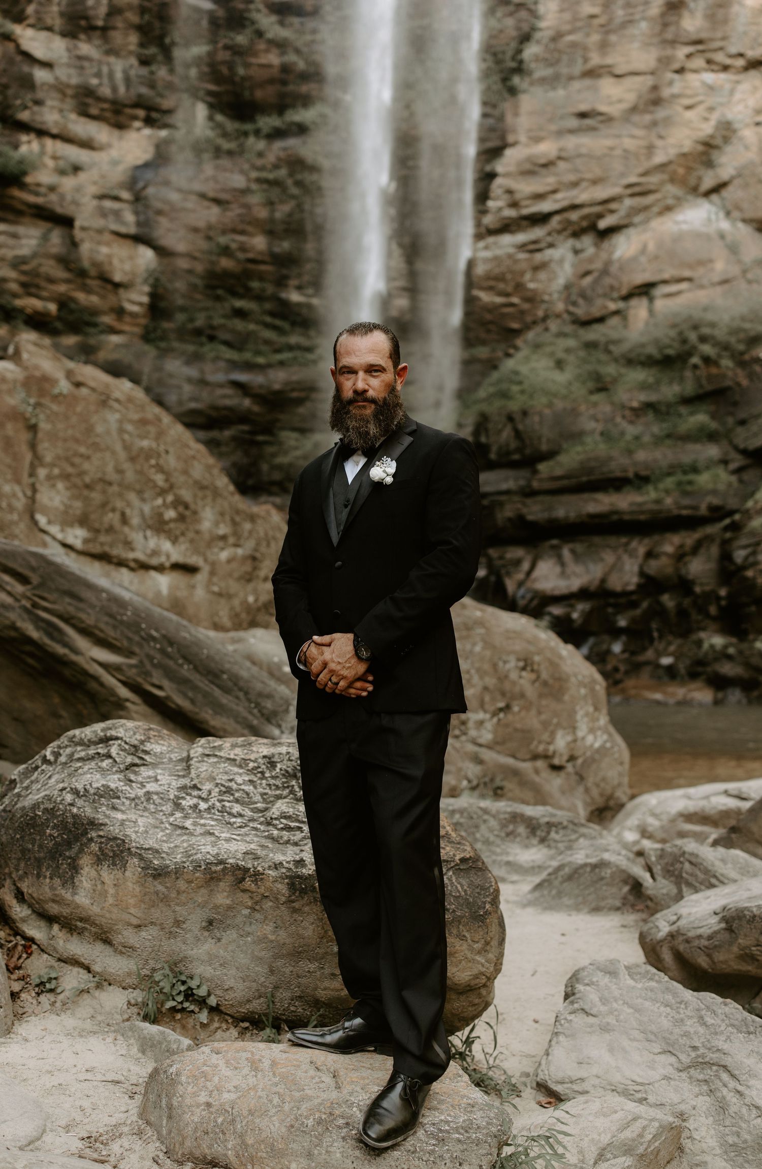Person in formal black attire stands pensively near rocky cliffs and waterfall in moody outdoor setting.