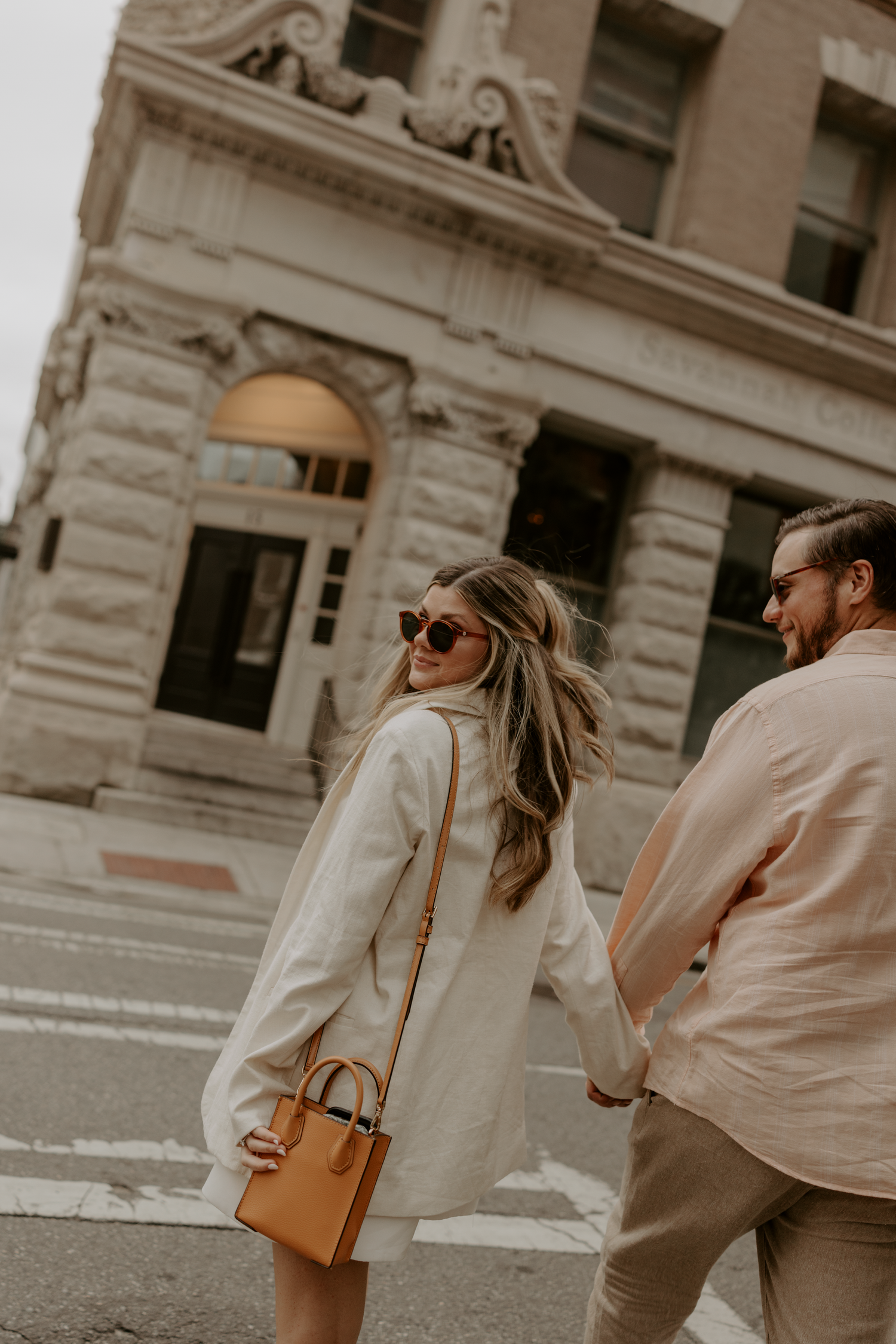Couple holding hands crossing a street, woman in cream coat with tan handbag and sunglasses, man in beige shirt.