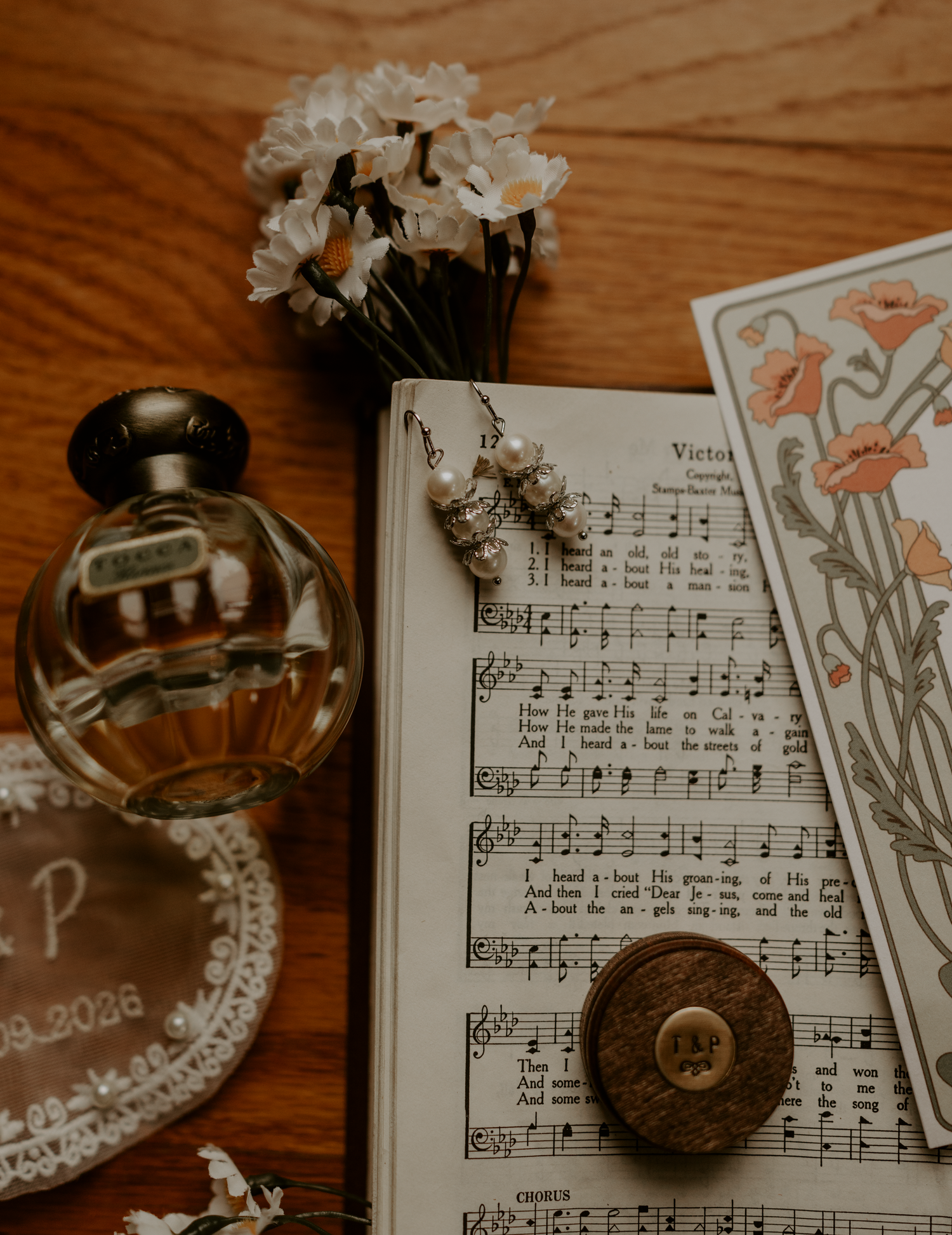 Vintage wedding flat lay with sheet music, perfume bottle, earrings, engraved dish, floral card, and white flowers on wood.