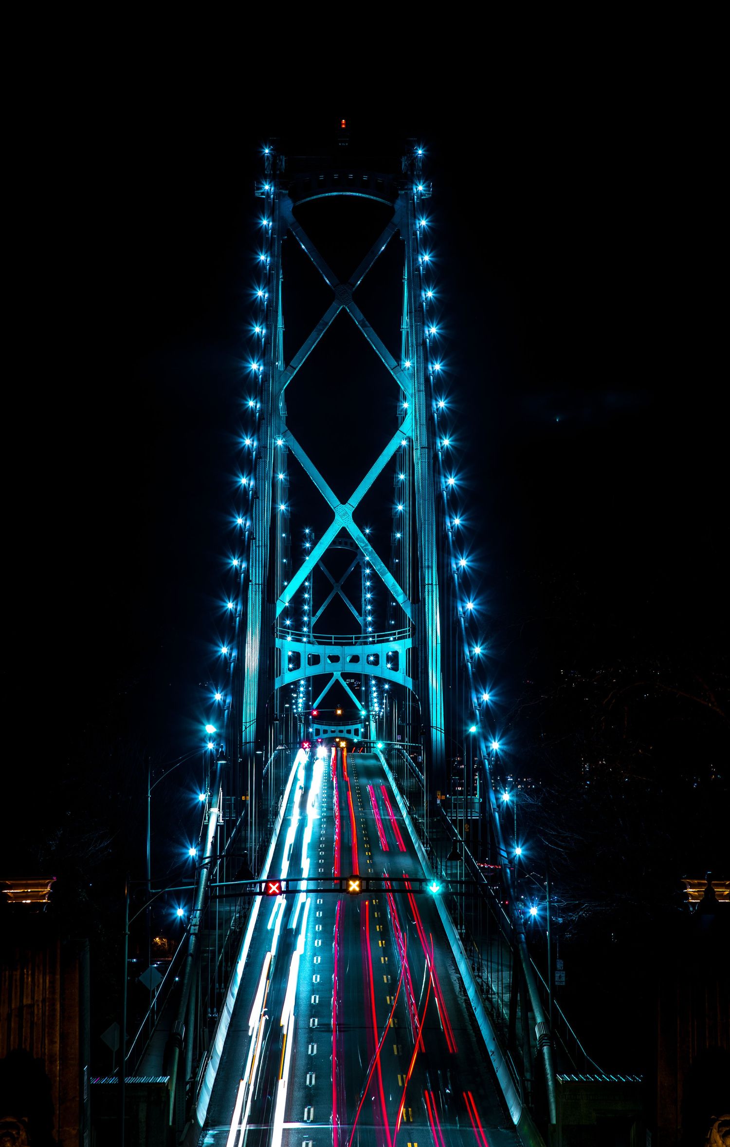 Lions Gate Bridge illuminated with blue lights reflects traffic trails at night.