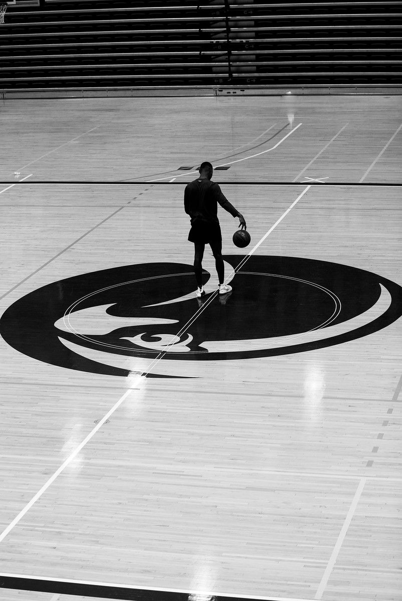 A silhouetted basketball player dribbles on an empty indoor court with a large logo.