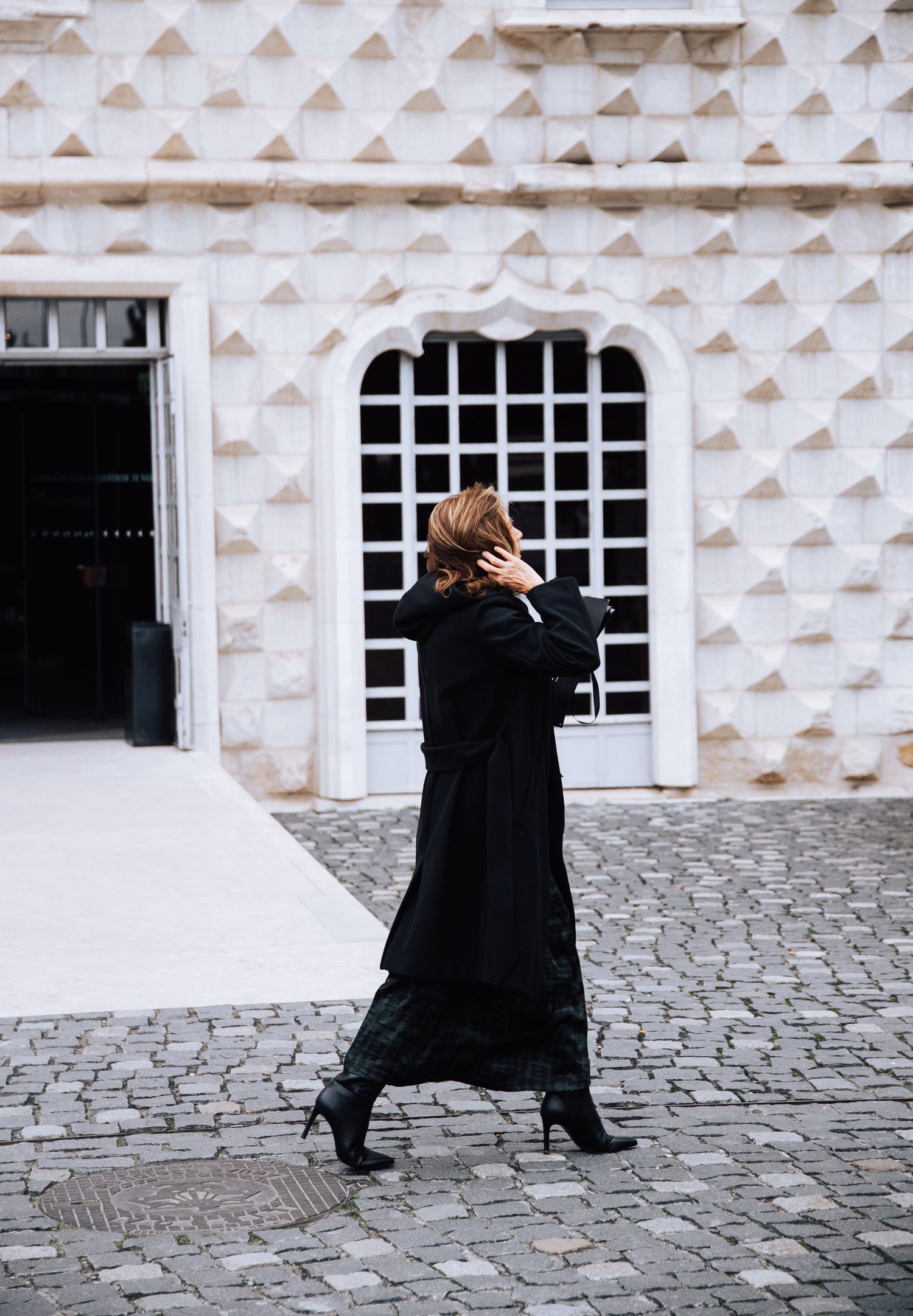 Figure in black coat walks against textured white building wall with diamond patterns. Lisbon.