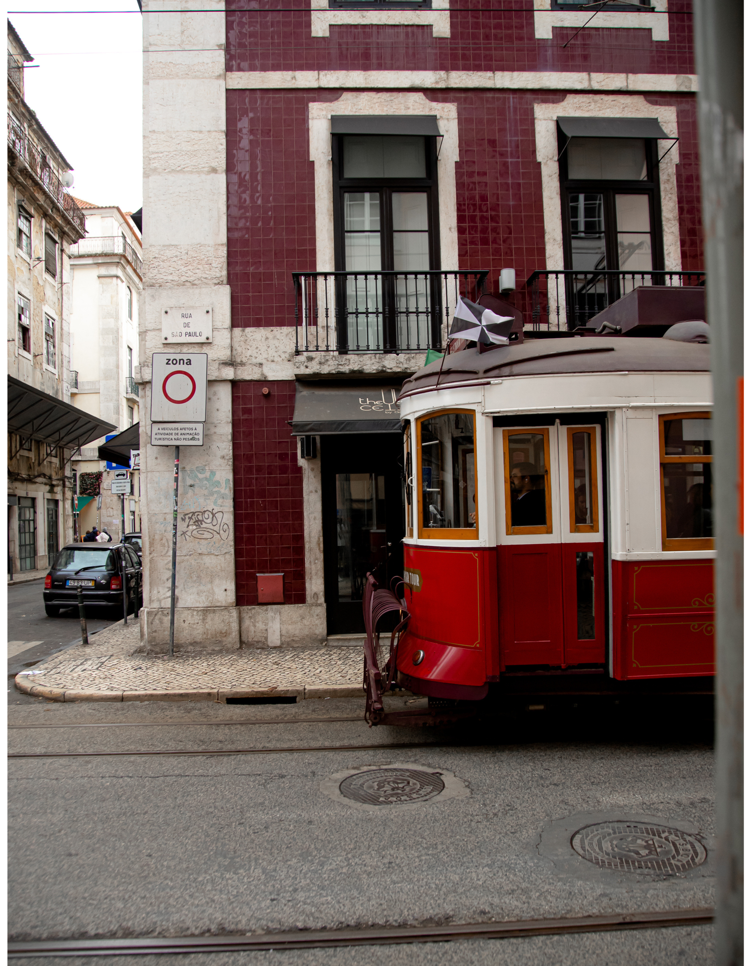 A vintage red tram navigates through narrow streets of an old European city with burgundy buildings.