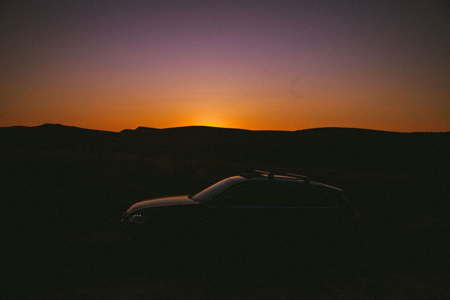 Silhouette of a car against dramatic sunset sky over mountain landscape.