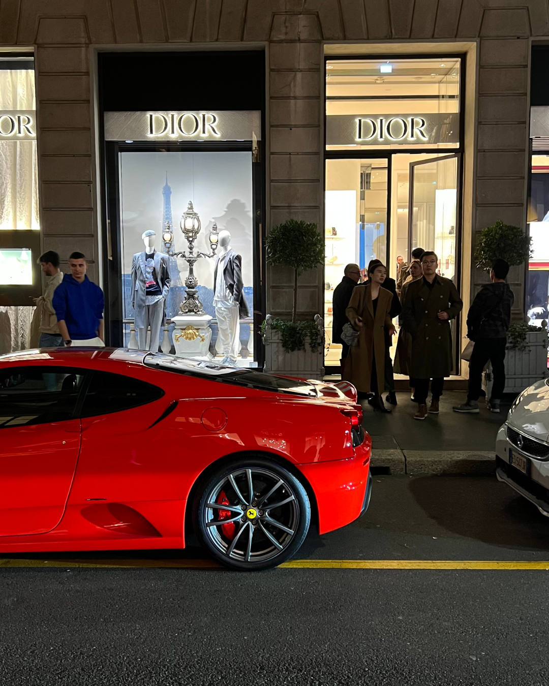 Red Ferrari parked outside illuminated Dior storefront at night. Milan, Italy.