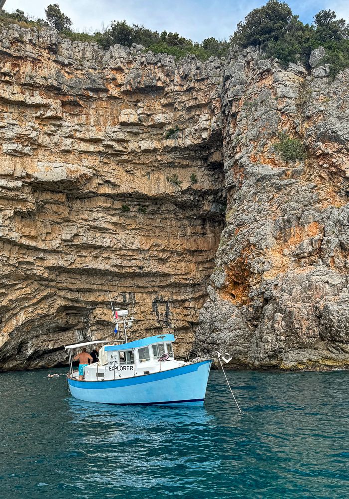 Bright blue tourist boat anchored near dramatic limestone cliffs along a Mediterranean coastline.