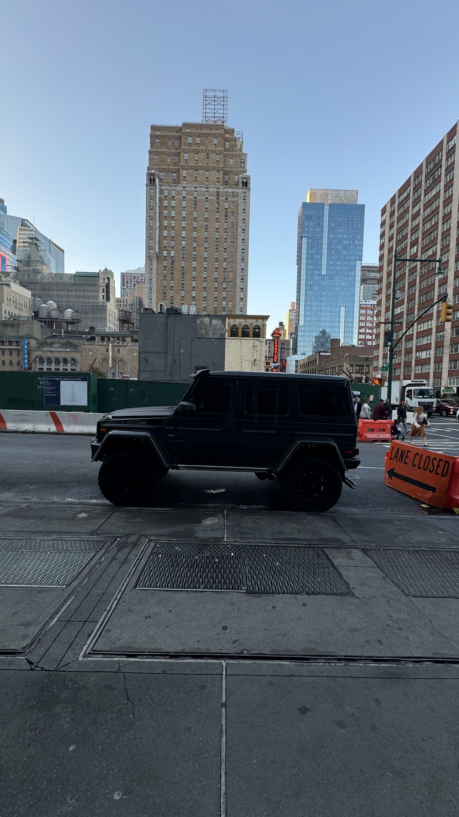 Black Mercedes G-Wagon parked in urban setting with tall skyscrapers and construction barriers visible.