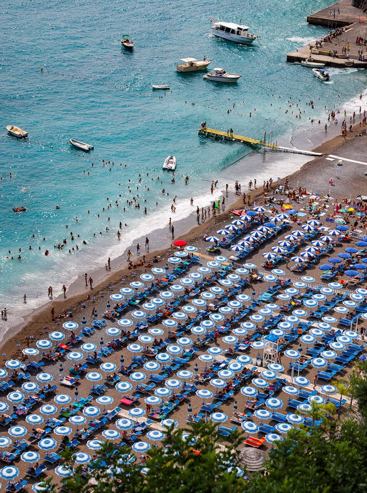 Aerial view of a crowded beach with organized rows of blue and white striped umbrellas and boats in the water.