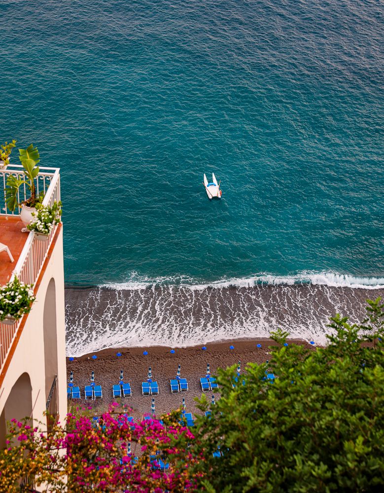 Colorful Mediterranean beach scene with blue umbrellas and flowering plants overlooking turquoise waters.