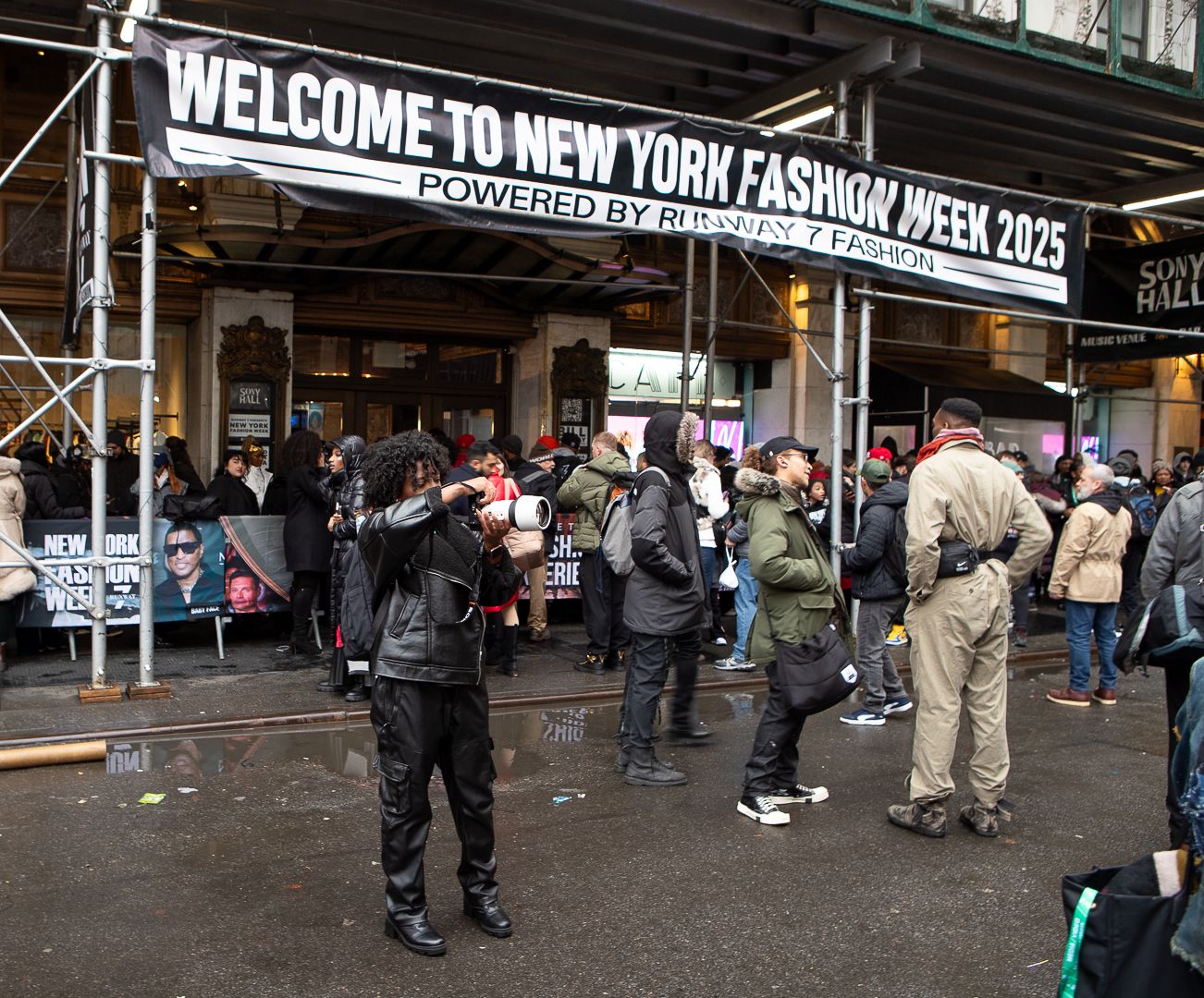 Crowd gathering under 'Welcome to New York Fashion Week' banner on a cloudy day.
