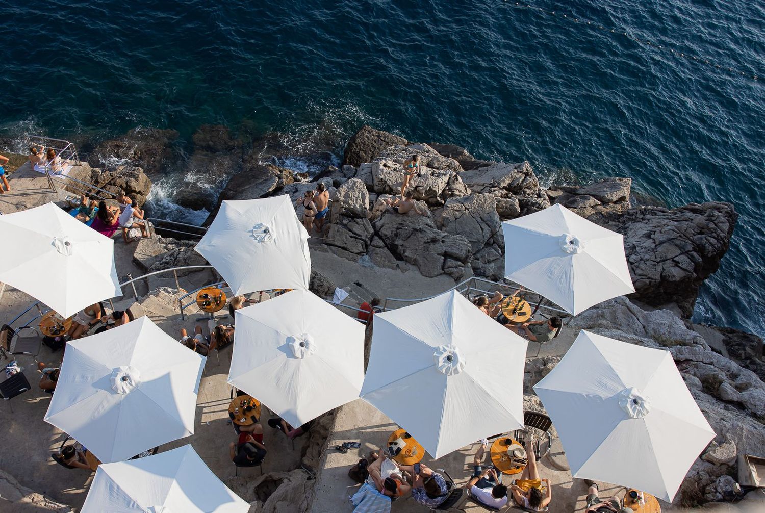 Aerial view of white umbrellas and seating along a rocky coastal edge.
