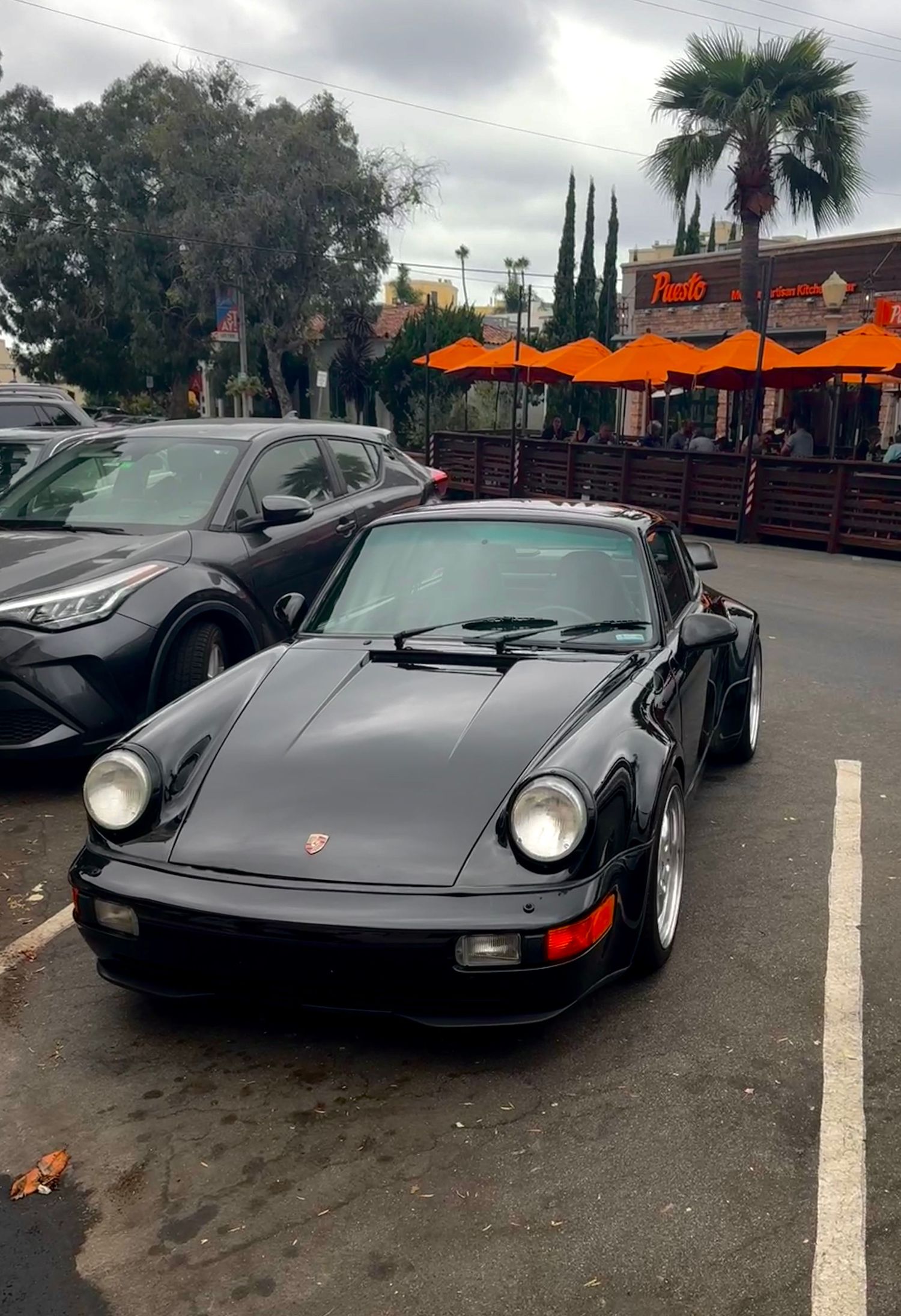 A black vintage Porsche 911 sits in a parking lot with orange umbrellas and palm trees visible in the background.