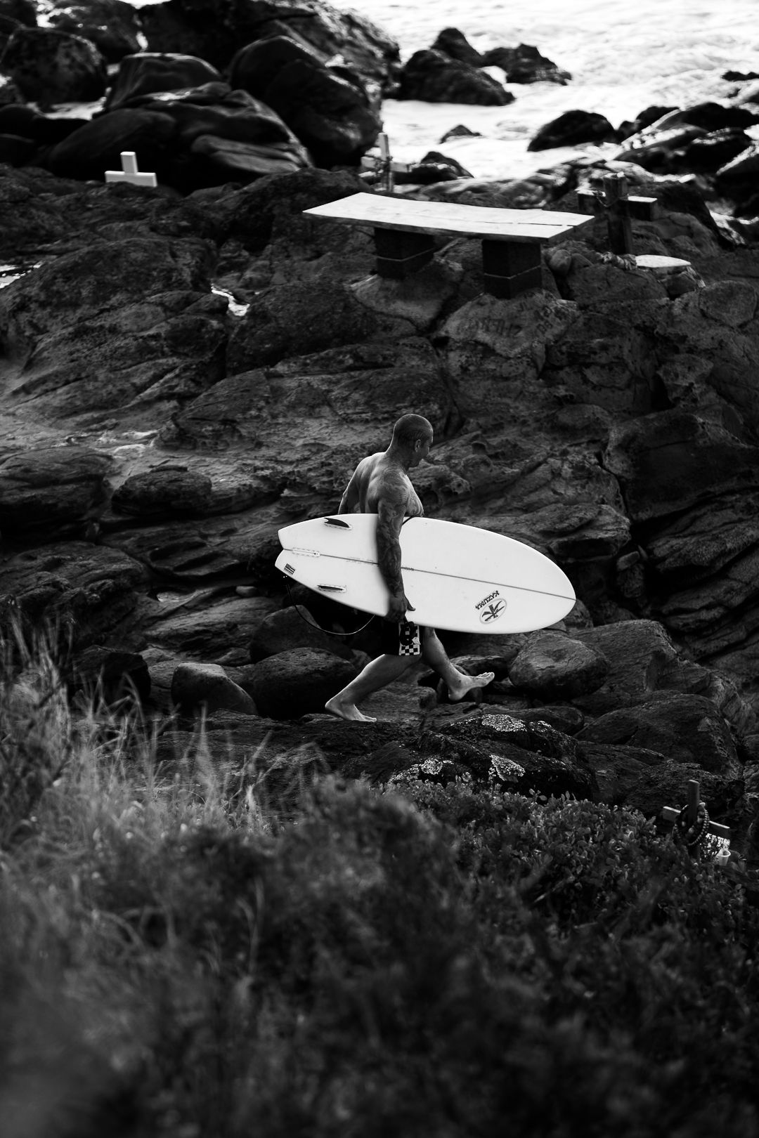 A surfer carrying a white surfboard navigates rocky coastal terrain in black and white. Maui, HI.