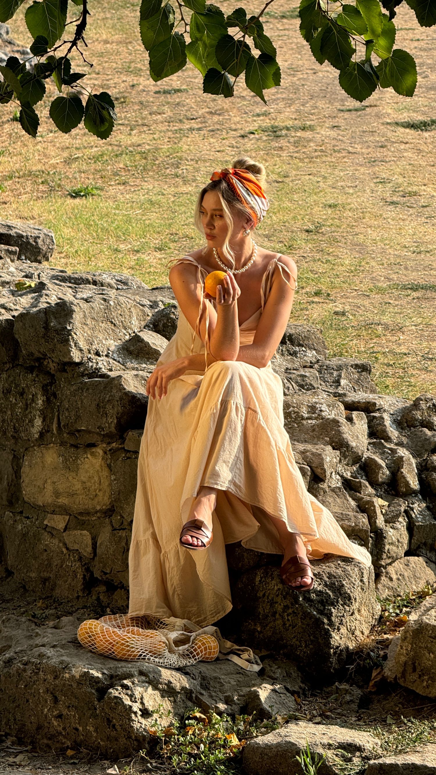 A person in a flowing yellow dress and headscarf sitting on stone wall in summer sunlight. Belgrade, Serbia.