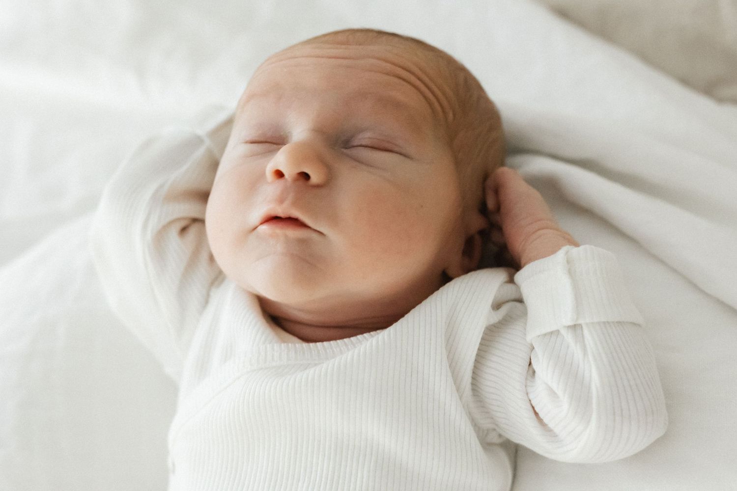 Rauhallisesti nukkuva vastasyntynyt vauva valkoisella vuodevaatteella, yllään valkoinen body ja käsi kohotettuna kasvojen lähelle.