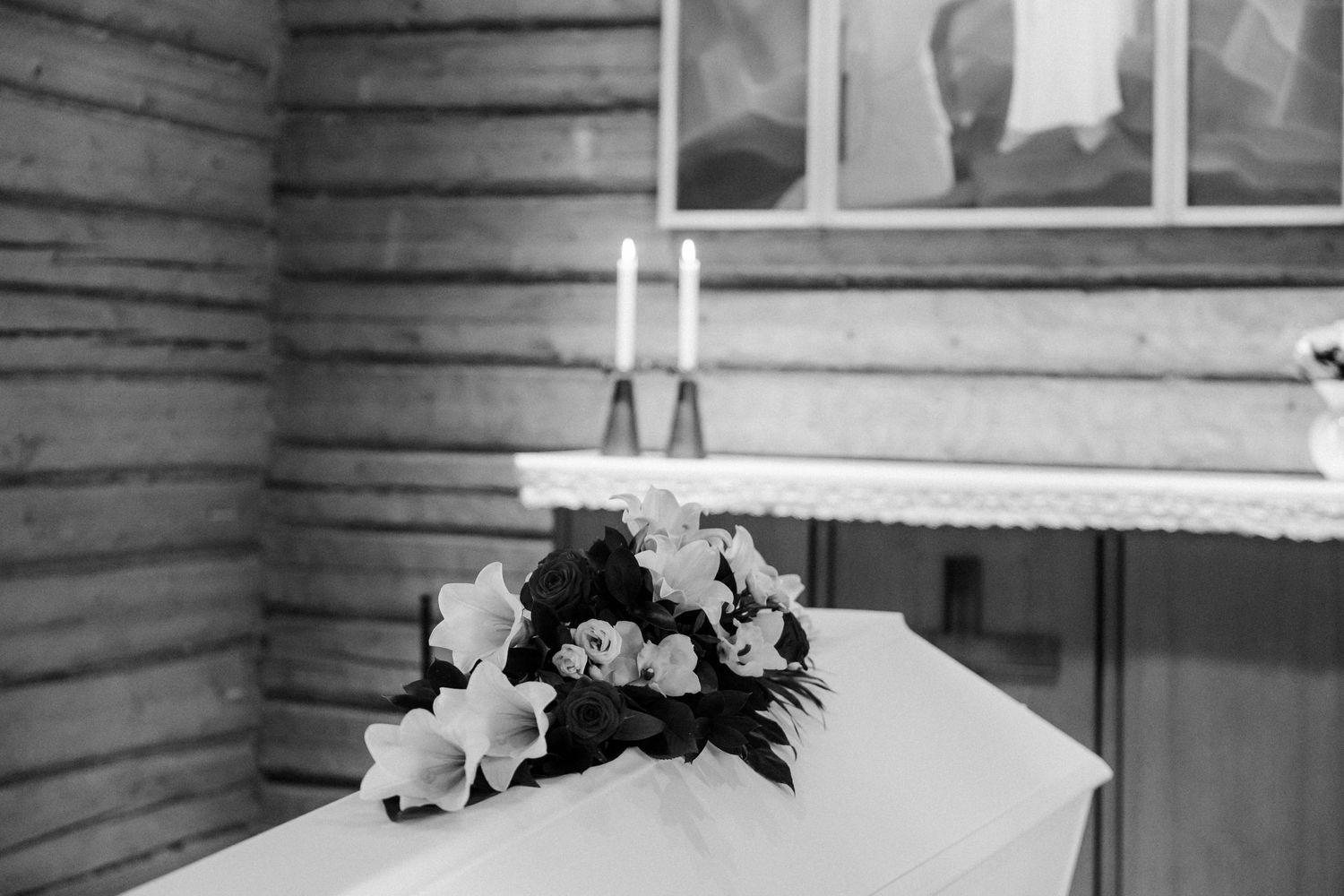 Mustavalkoinen kuva siunauskappelista. Valkoinen arkku, jonka päällä kukkalaite, taustalla kaksi kynttilää.