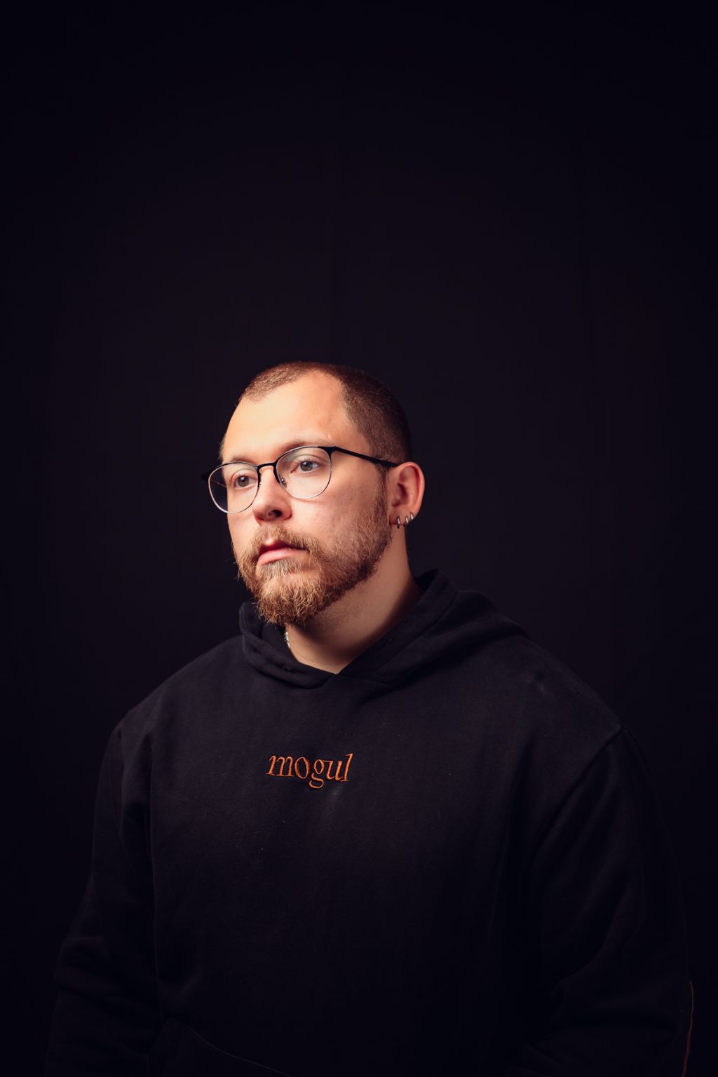 A bearded person wearing glasses and a black hoodie with MOGUL text poses against a dark background in a professional photo series.