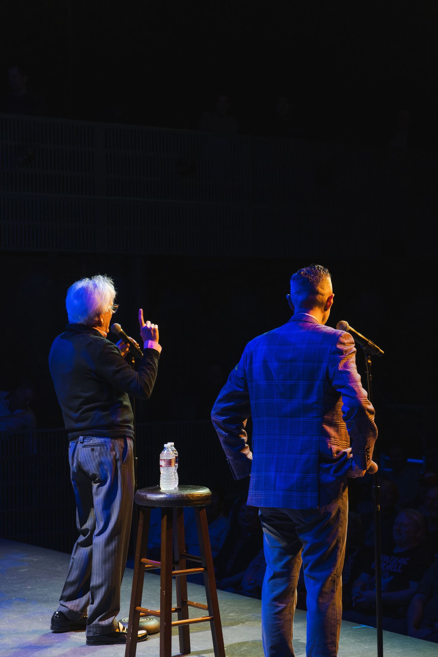 Two performers on stage with microphones in a darkened theater setting.