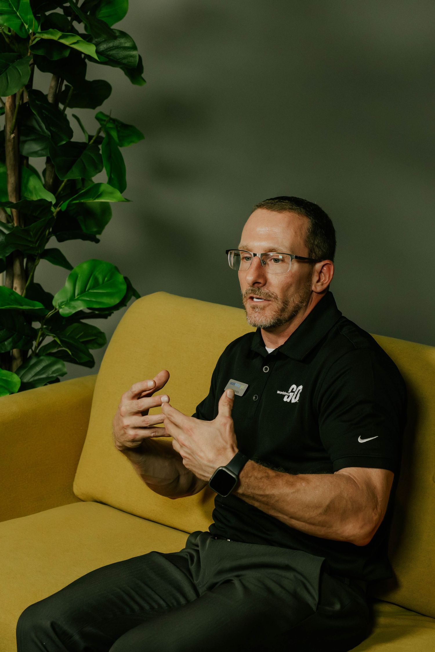 Person gesturing while sitting on yellow couch with plants in background.