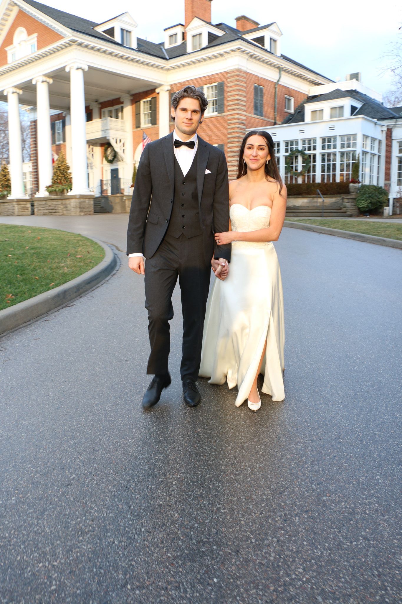 Wedding couple walking along a driveway in front of a grand colonial-style mansion.