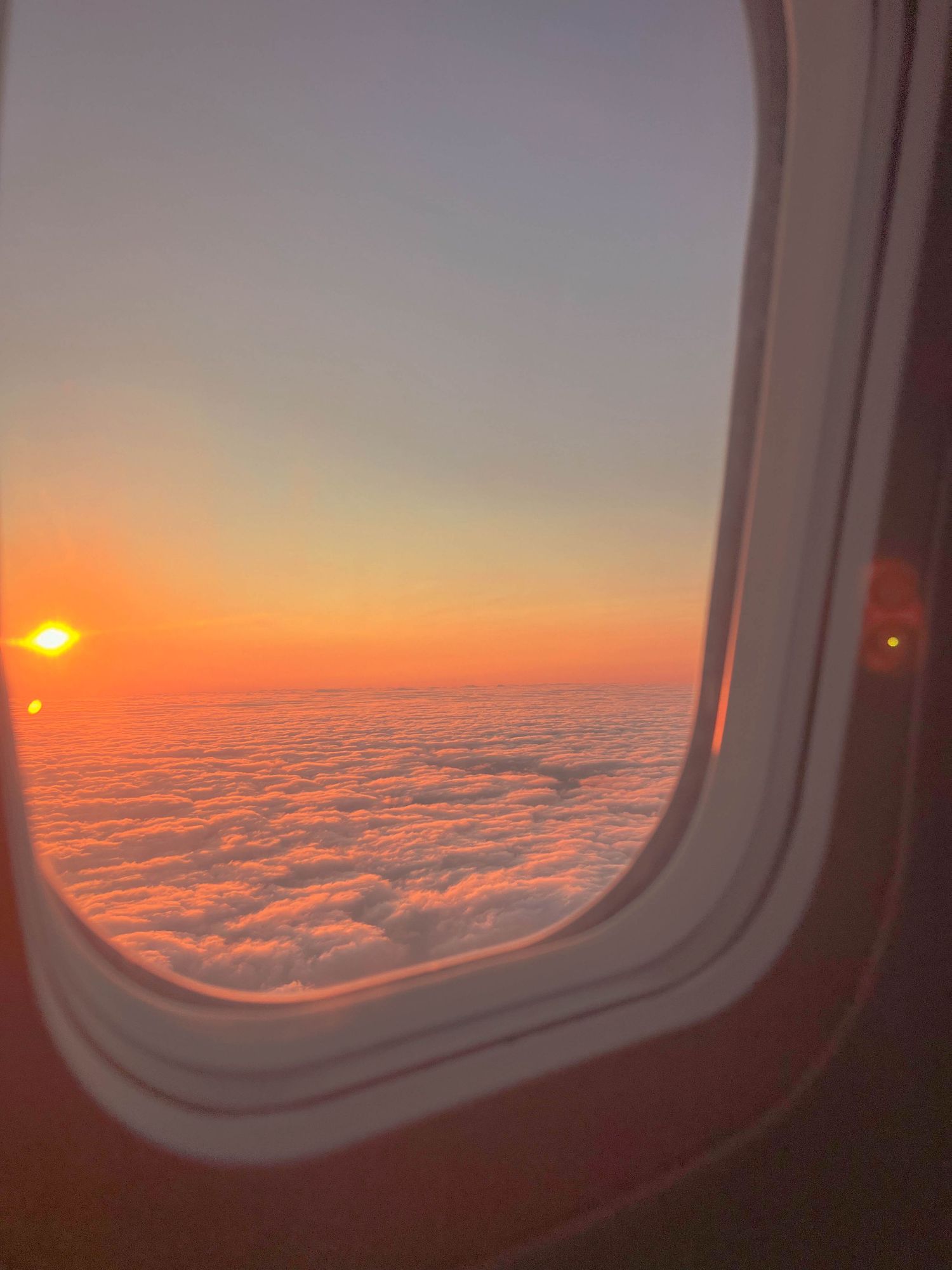 Sunset view from airplane window showing orange sun over sea of clouds.
