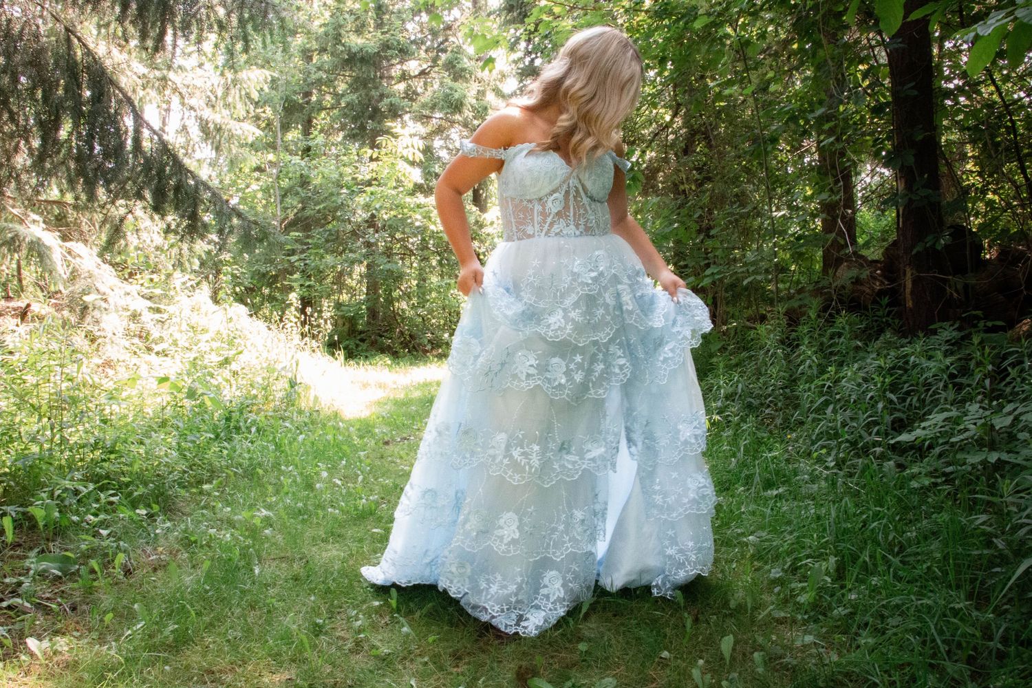 A bride in a lace wedding dress walks through a sunlit wooded forest path during a photoshoot.