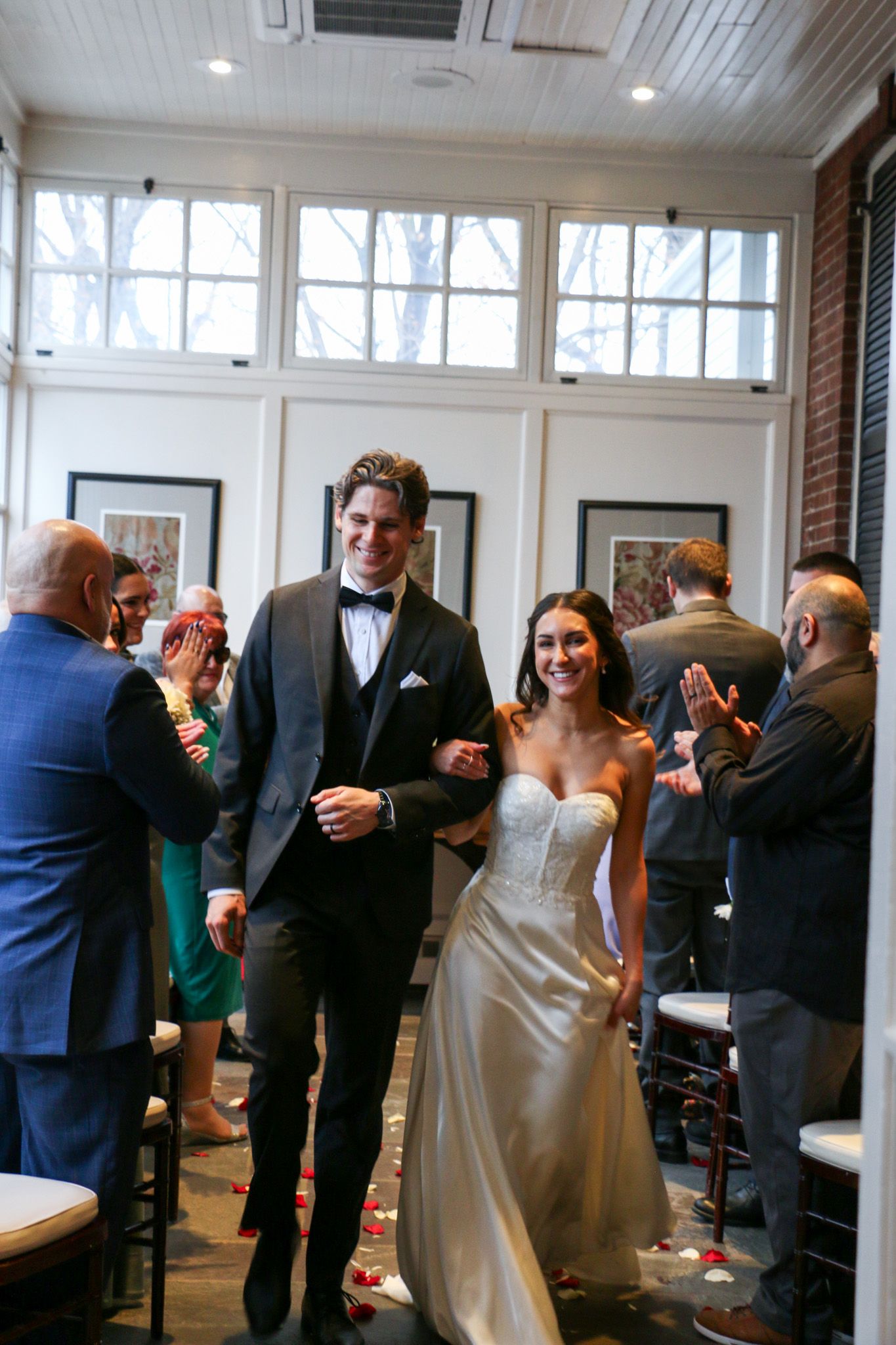 A newlywed couple walks down an aisle while guests celebrate at an indoor wedding reception venue.