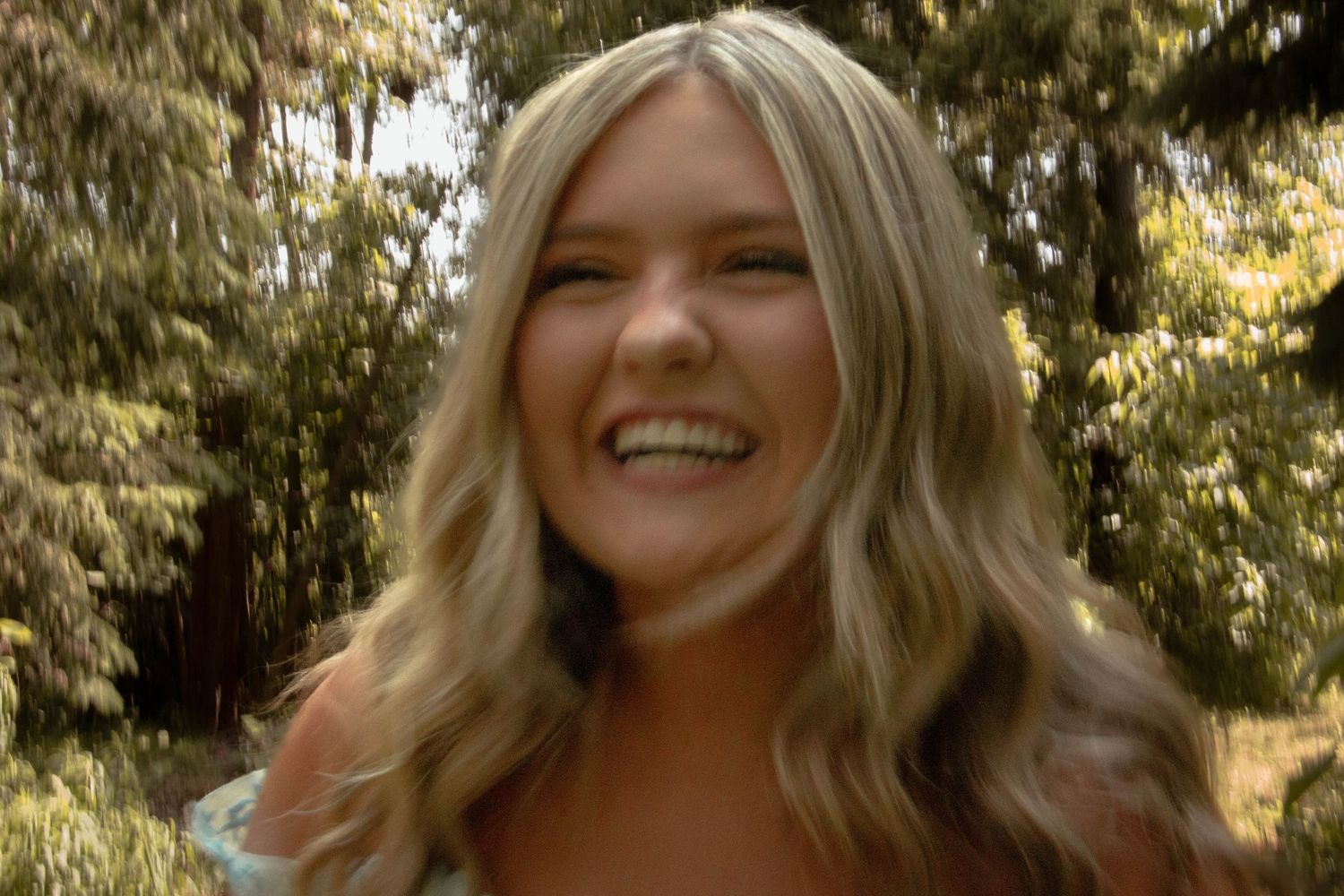 A person with long wavy blonde hair smiles warmly while standing outdoors in natural lighting.