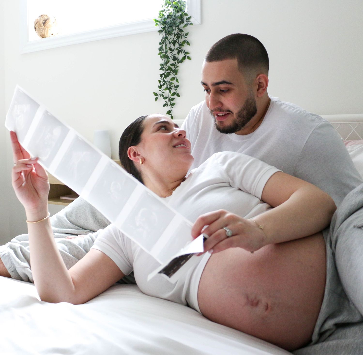 Couple relaxes on white bedding while reviewing pregnancy ultrasound images.